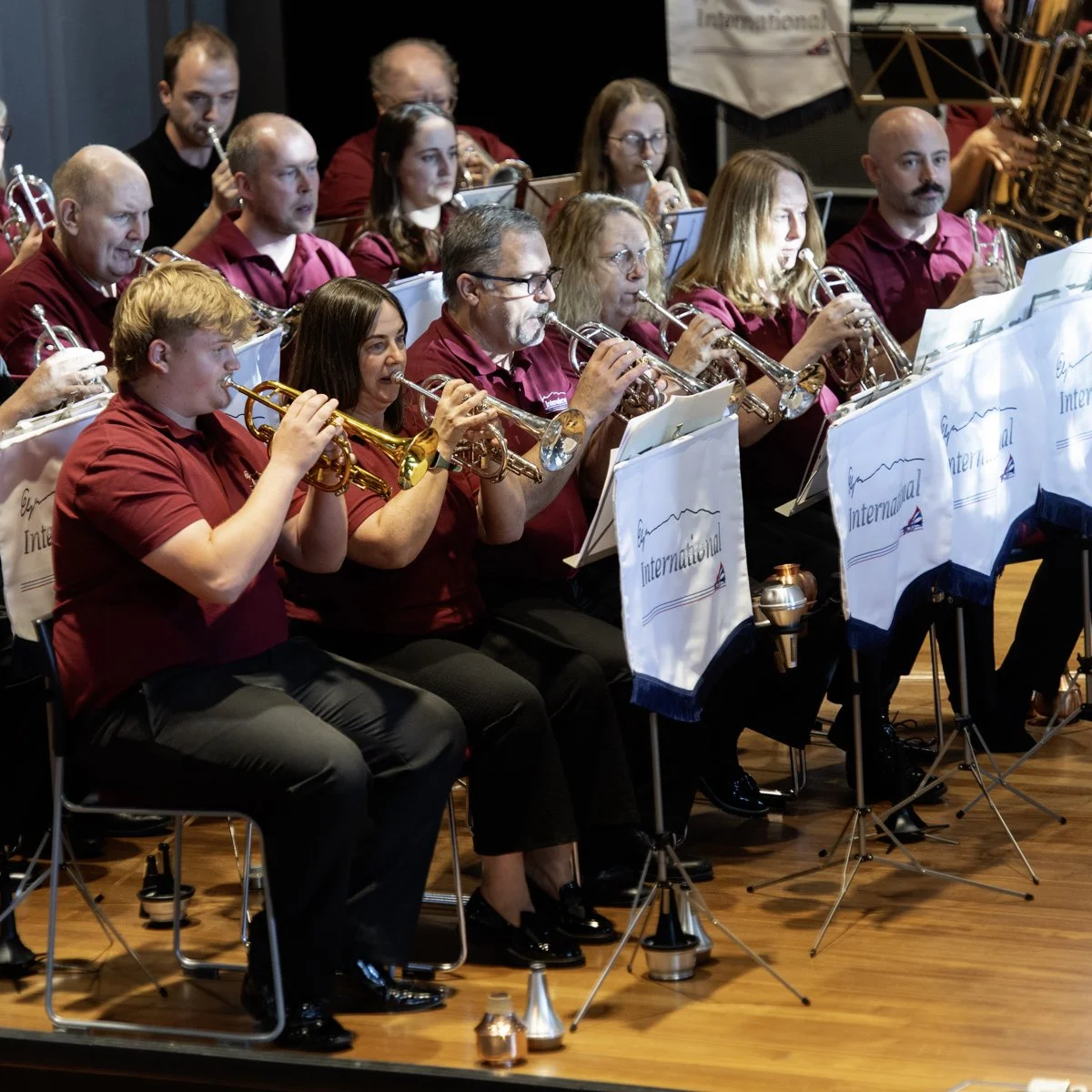 A group of musicians playing trumpets during a performance, seated behind music stands with banners reading "International." They are wearing matching maroon shirts, and the setting appears to be an indoor concert hall.