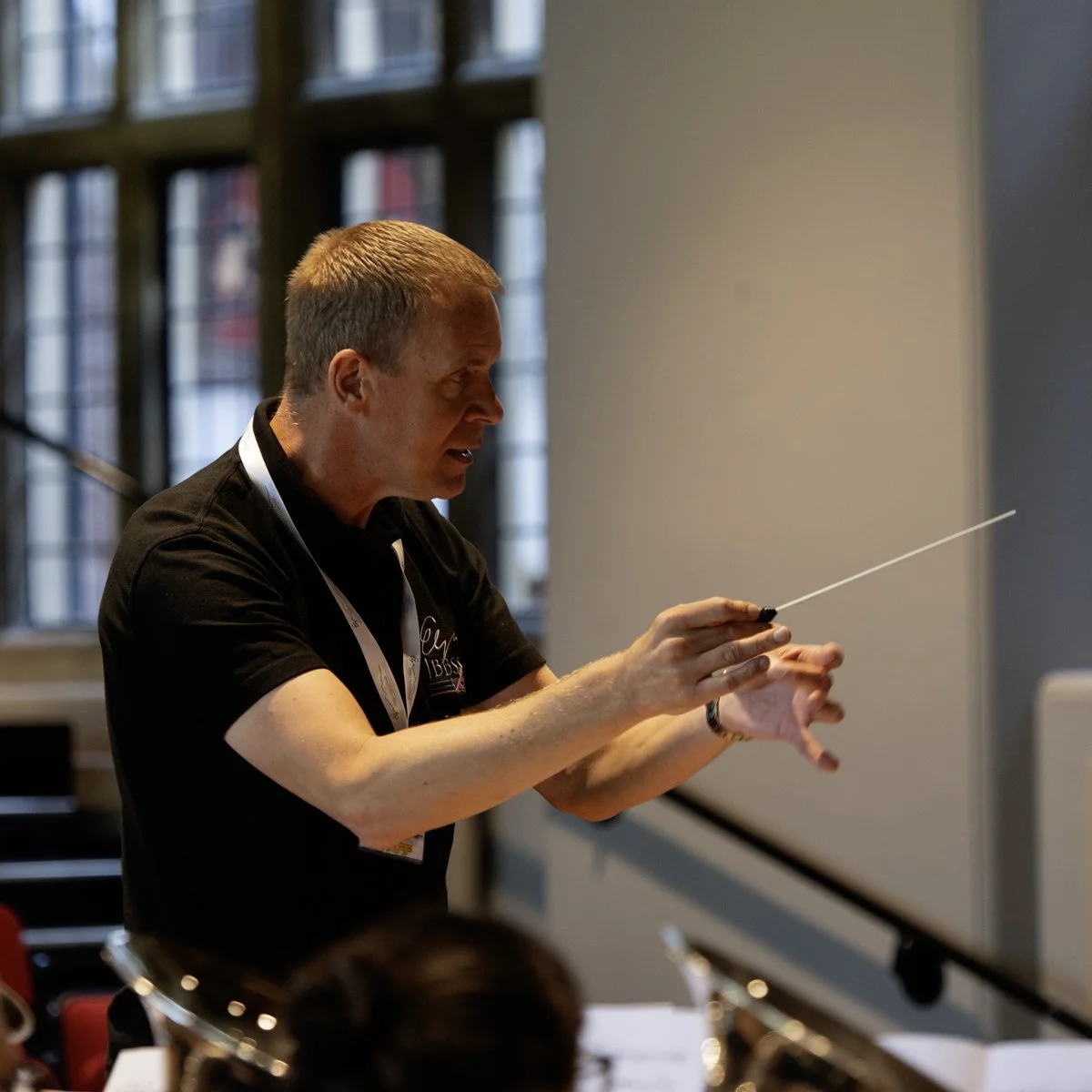 A man conducting an orchestra with a baton in front of a music stand inside a building with large windows.