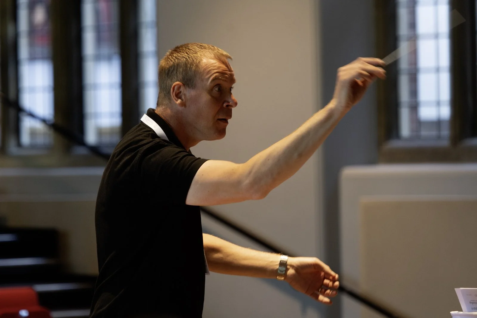 A man conducting or gesturing with his right hand during a performance or presentation inside a room with tall windows.