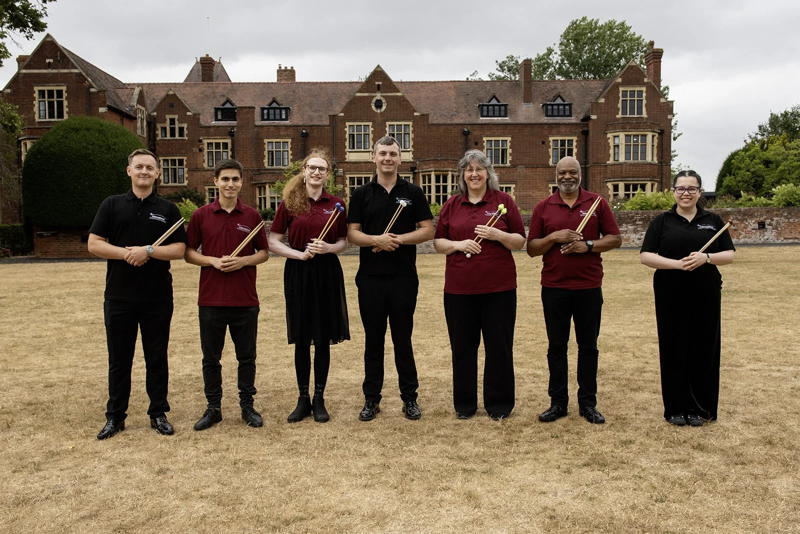 Group of seven people standing outdoors on a lawn in front of a large brick building, holding drumsticks, smiling