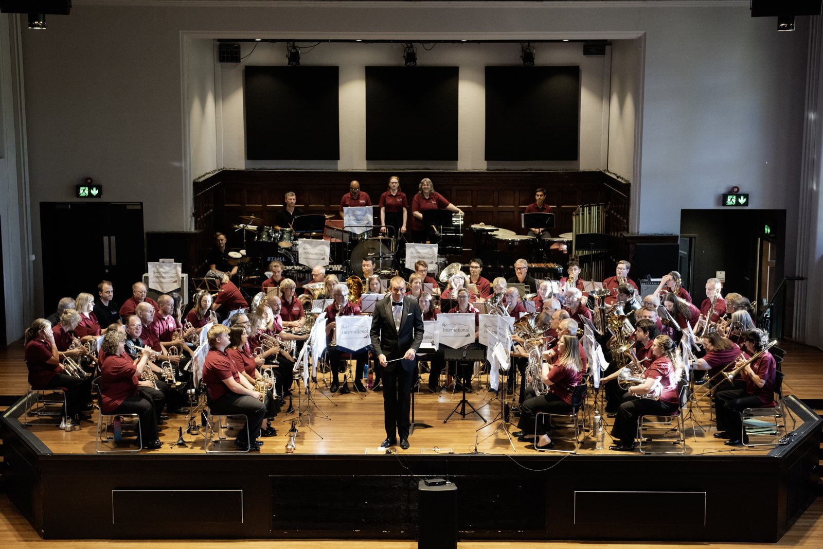 A large orchestra performing on a stage with a conductor in front. The musicians are seated in rows playing various instruments, including brass, woodwinds, and percussion. The stage has a wooden floor and is set in a concert hall with black backdrop