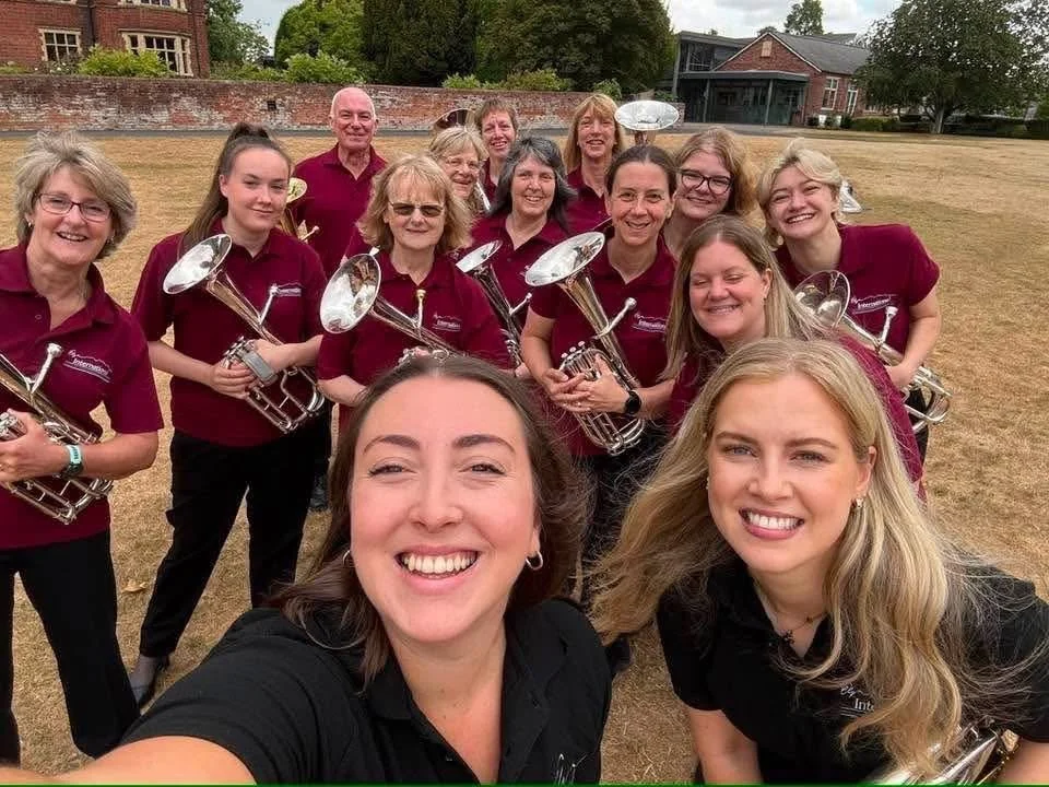 Group of women and one man, part of a marching band, standing outdoors on a grassy field, holding trumpets, smiling for a selfie.