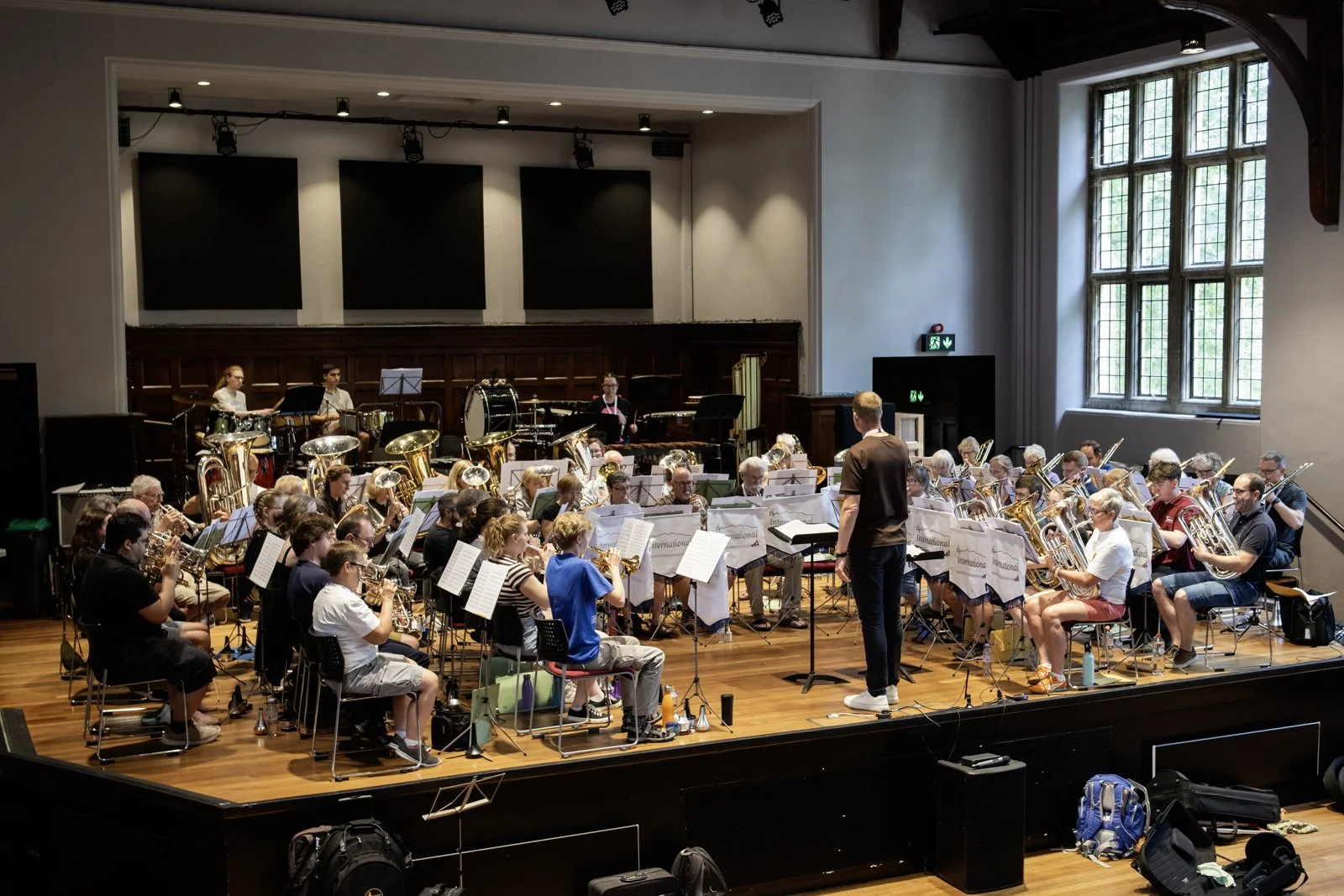 Large orchestral ensemble rehearsing in a spacious concert hall with a conductor and multiple musicians playing instruments, including brass, woodwinds, and percussion, in front of a wooden stage with large windows providing natural light.