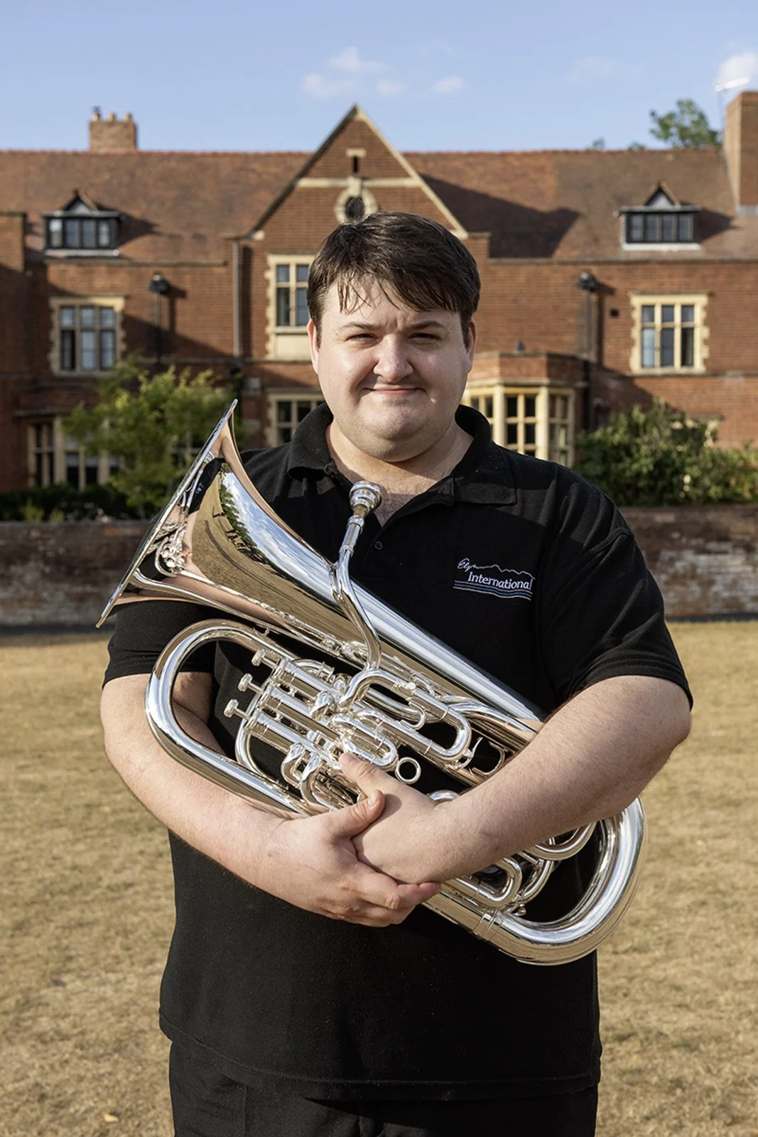 A man holding a silver euphonium in front of a large brick house with a red-tiled roof.