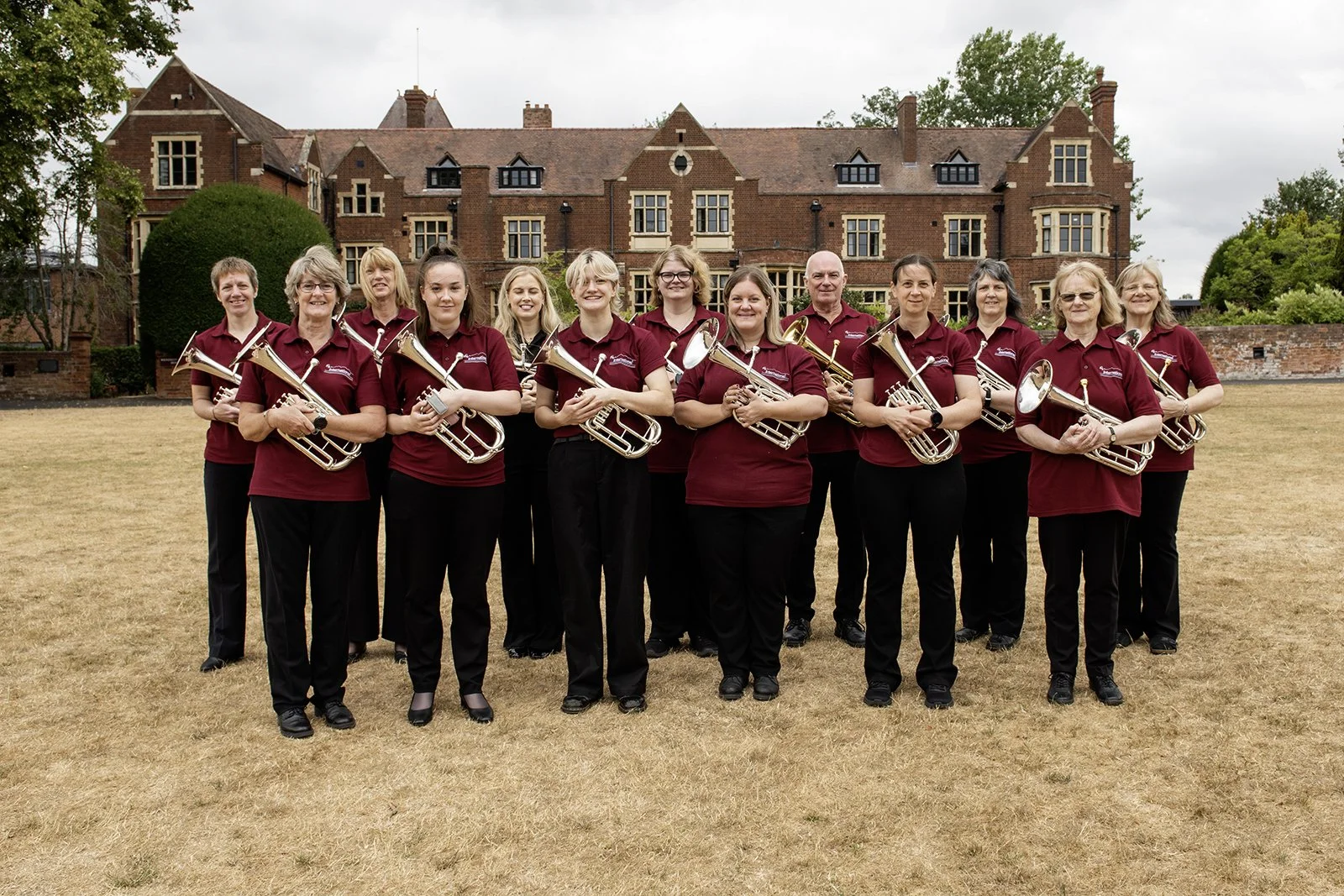 A group of thirteen women in matching maroon shirts and black pants standing outdoors on a dry grassy field, holding brass musical instruments, with a large brick building and trees in the background.