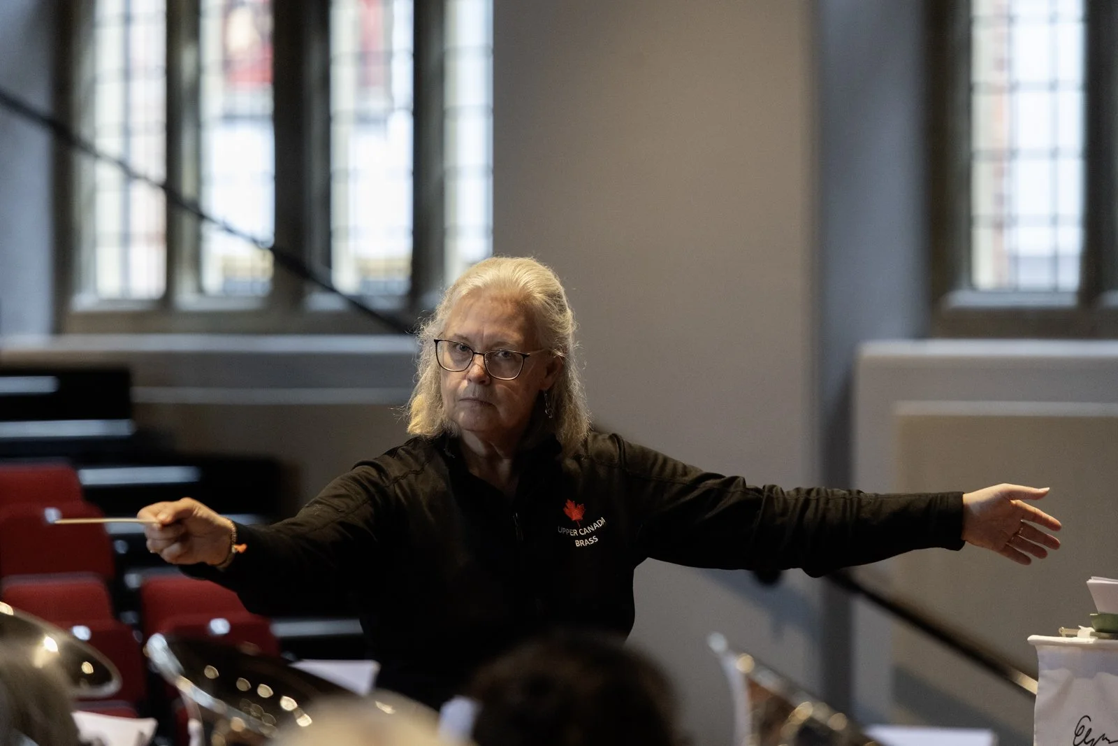 An older woman with glasses conducting an orchestra, wearing a black jacket with a red maple leaf and text that says 'UPPER CANADA BRASS'. She has grey hair and is in a room with large windows and red chairs.