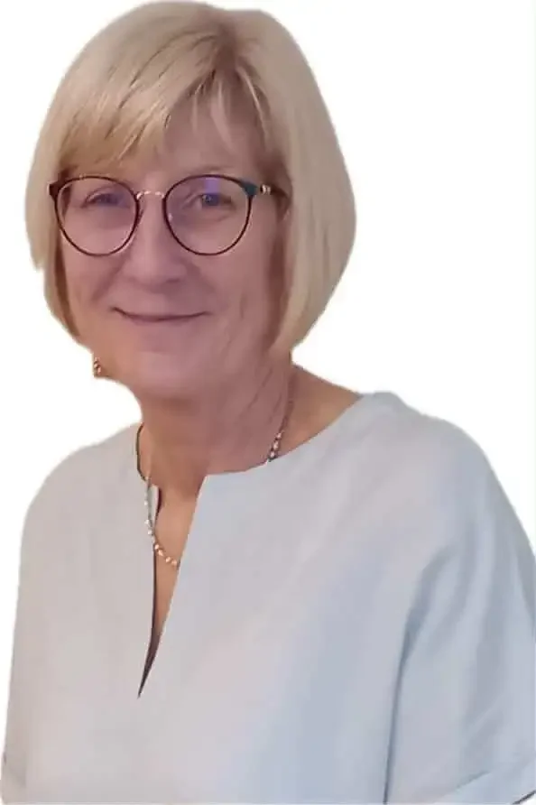A middle-aged woman with short blonde hair, glasses with round frames, and a light-colored blouse, smiling at the camera against a white background.