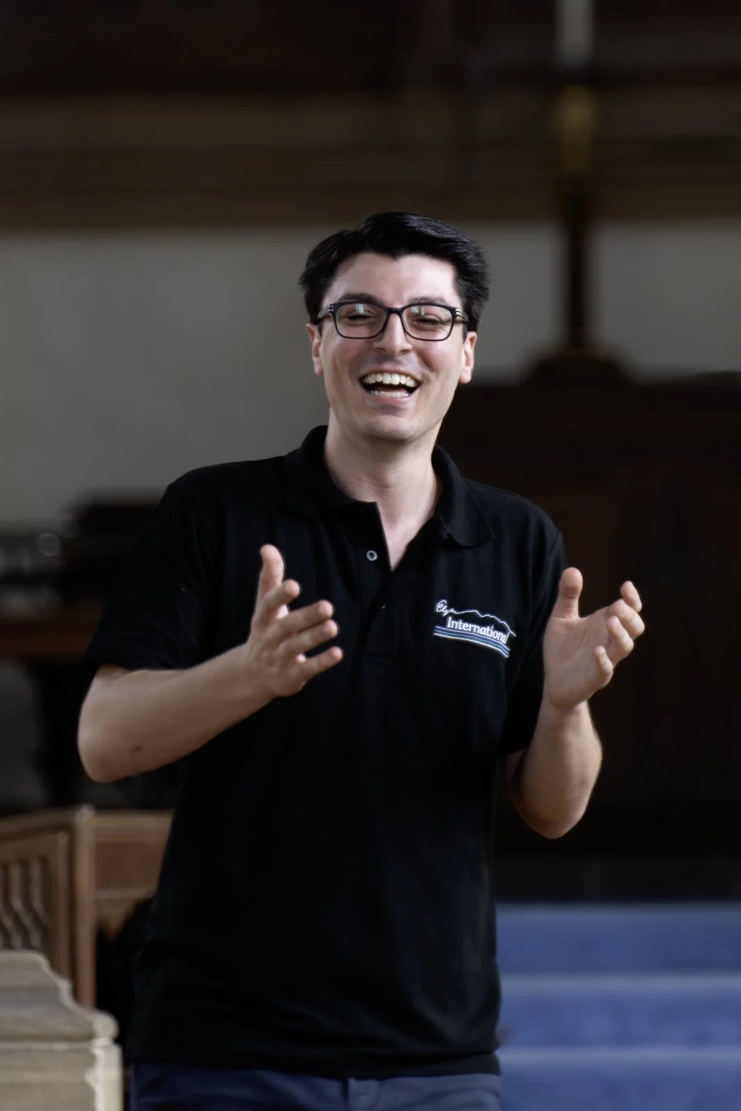 A man with glasses and dark hair, wearing a black polo shirt with a logo, is smiling and gesturing with his hands.