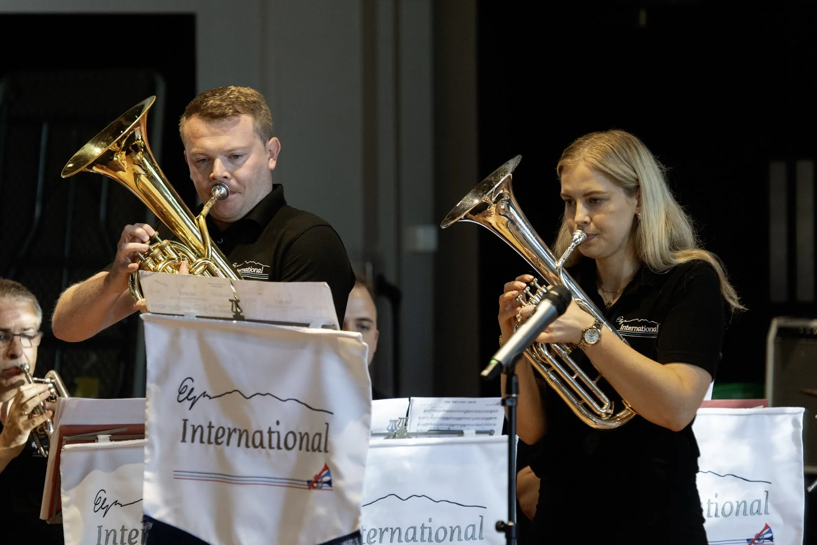 Two musicians playing brass instruments during a performance, with music sheets on music stands labeled 'International' in front of them.