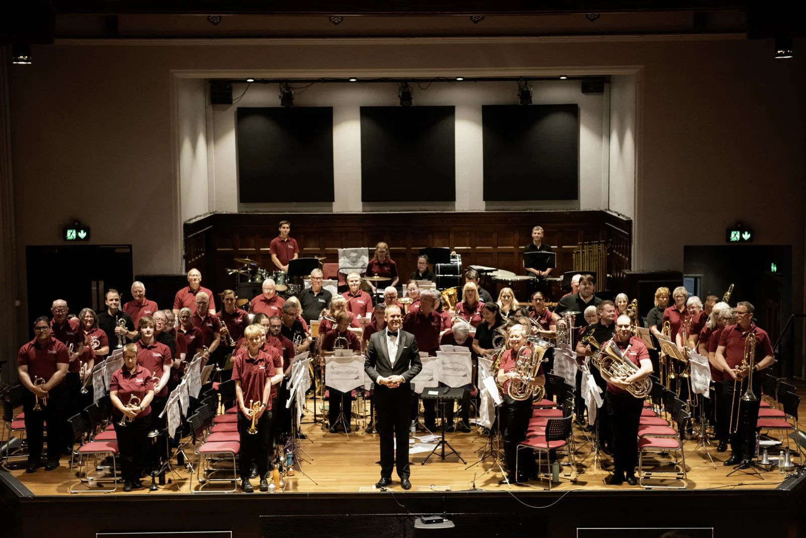 An orchestra of musicians standing on a stage in a concert hall, holding brass and percussion instruments, with a conductor in a tuxedo at the front. The hall has a wooden floor and three large black panels hanging on the back wall.