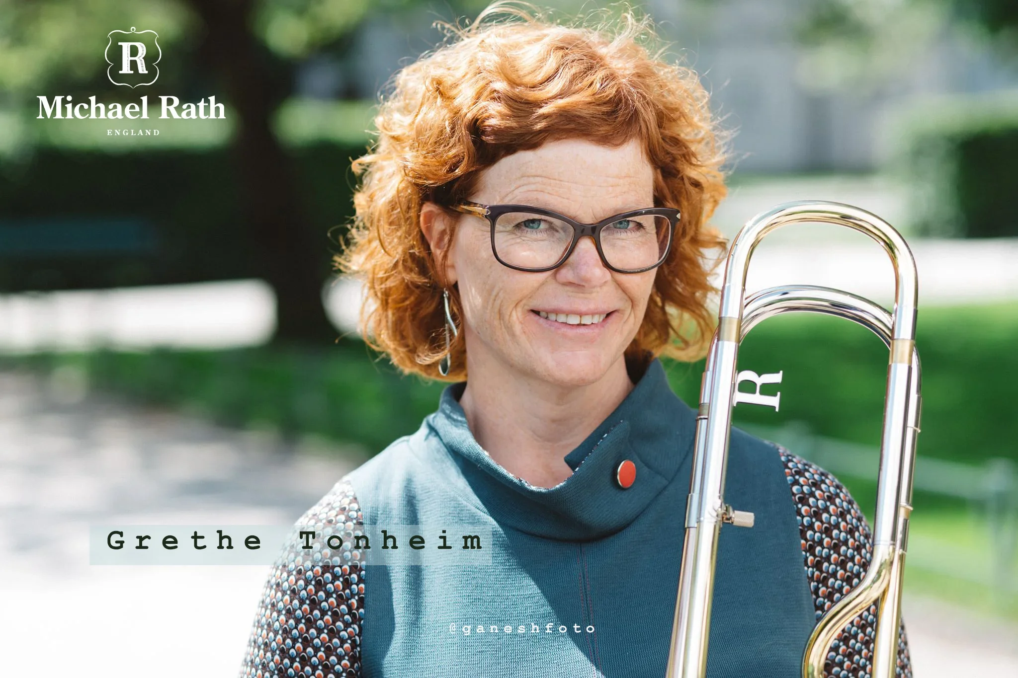 A woman with curly red hair, glasses, and a blue jacket smiling outdoors, holding a trombone, with green trees in the background.