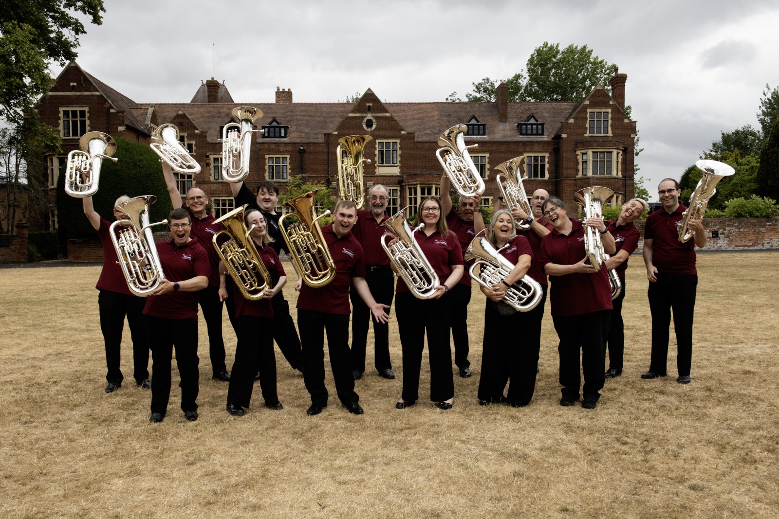 A group of people in matching maroon shirts holding up brass musical instruments, smiling and posing outdoors on a dry lawn with a large historic brick building in the background.