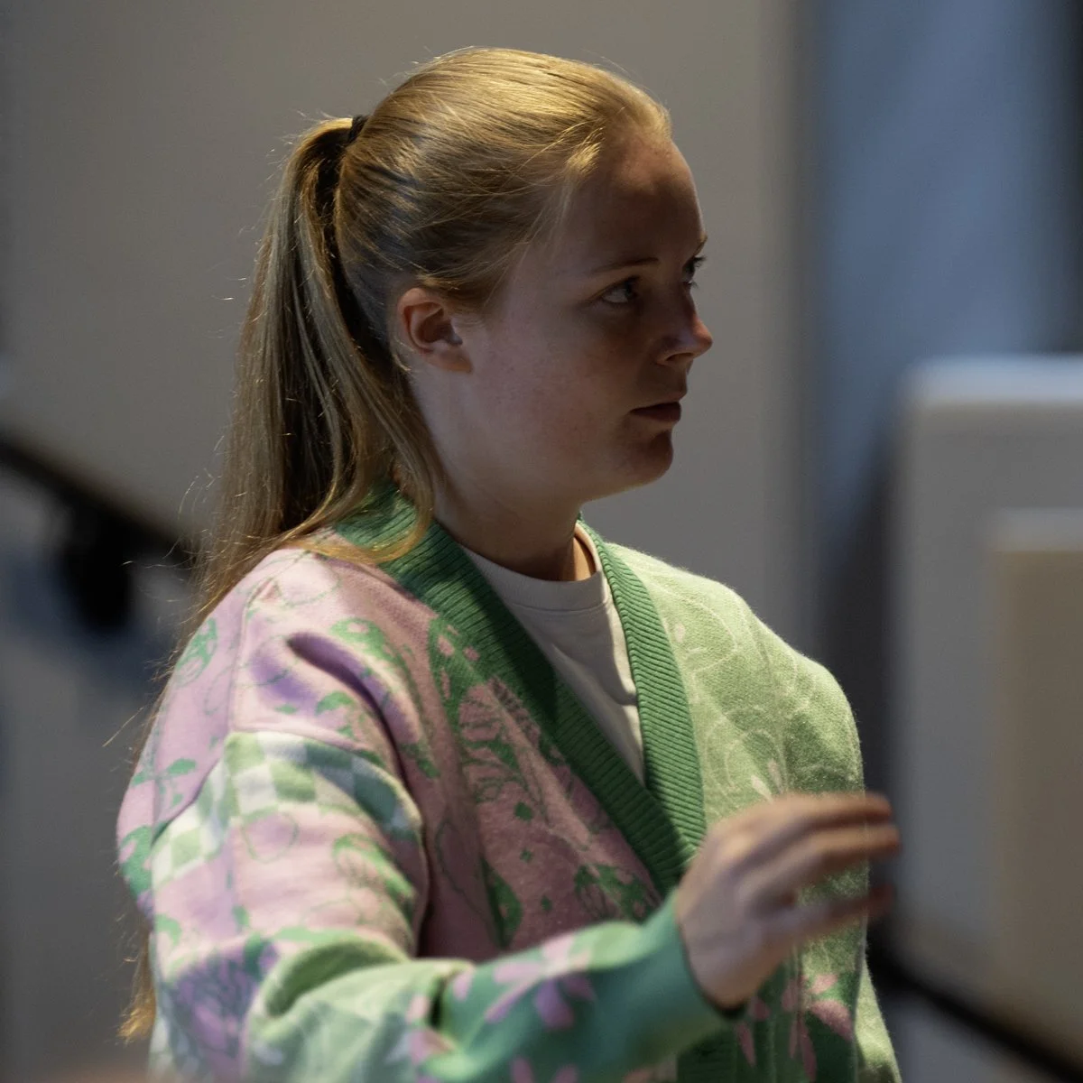 Side profile of a young woman with blonde hair in a ponytail, wearing a pastel pink and green patterned sweater with a white shirt underneath.