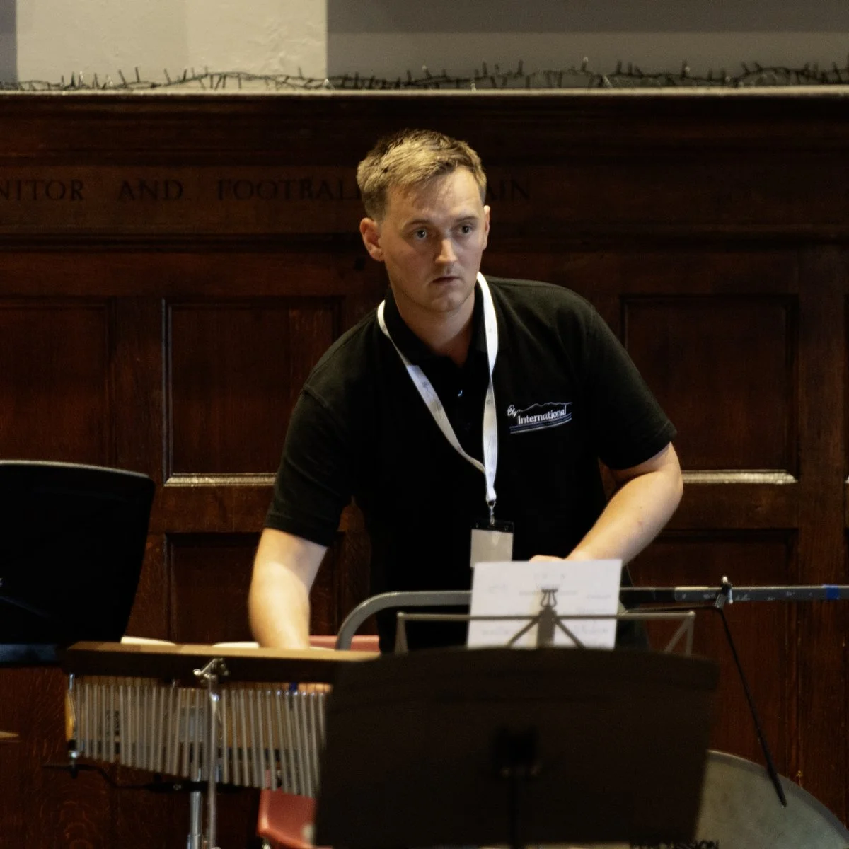 A young man with light brown hair playing a percussion instrument, possibly a vibraphone, in a room with wooden paneling, wearing a black shirt with a white lanyard and badge.