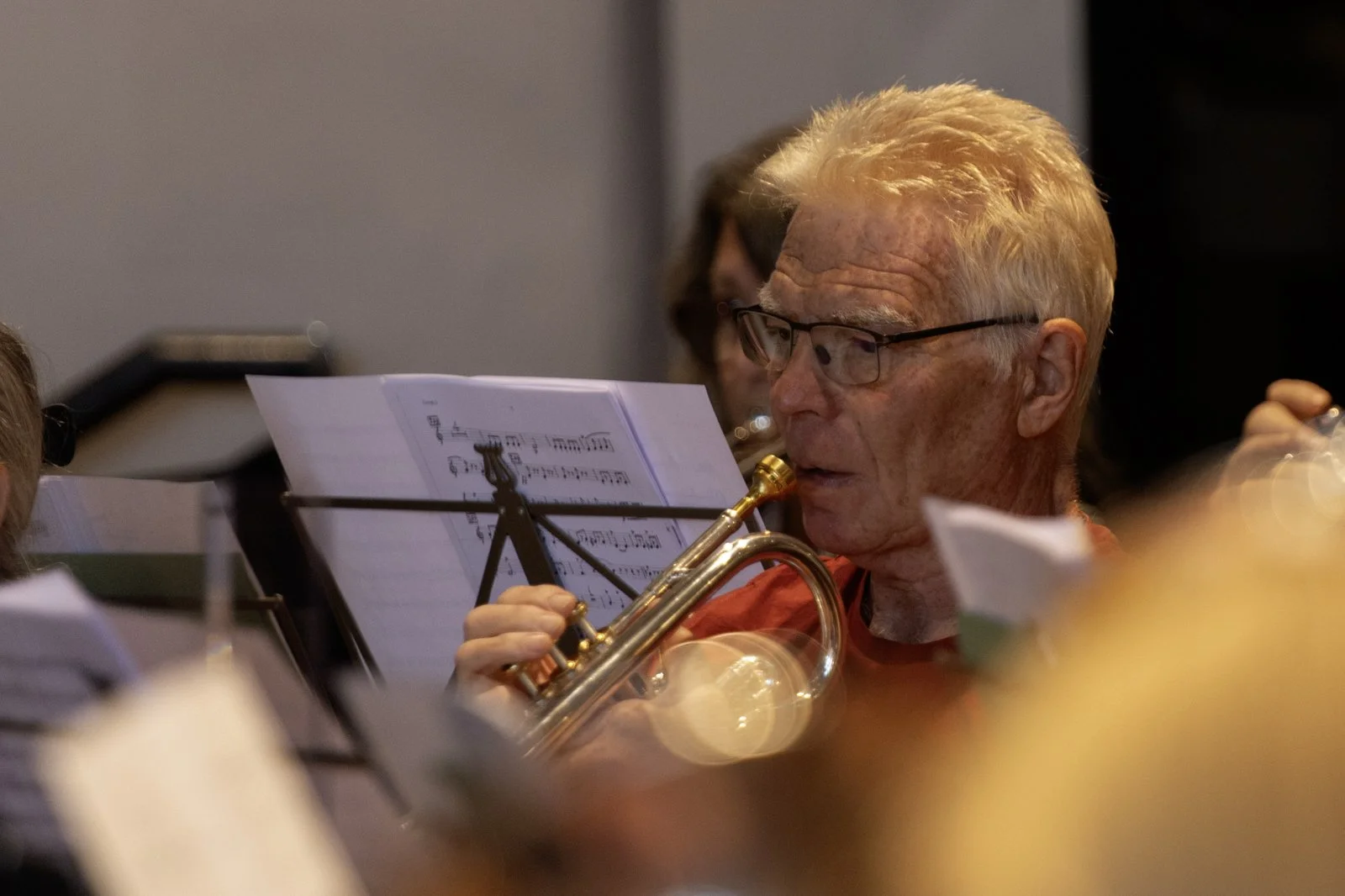 An elderly man with glasses playing a trumpet in an orchestra, surrounded by sheet music and other musicians.