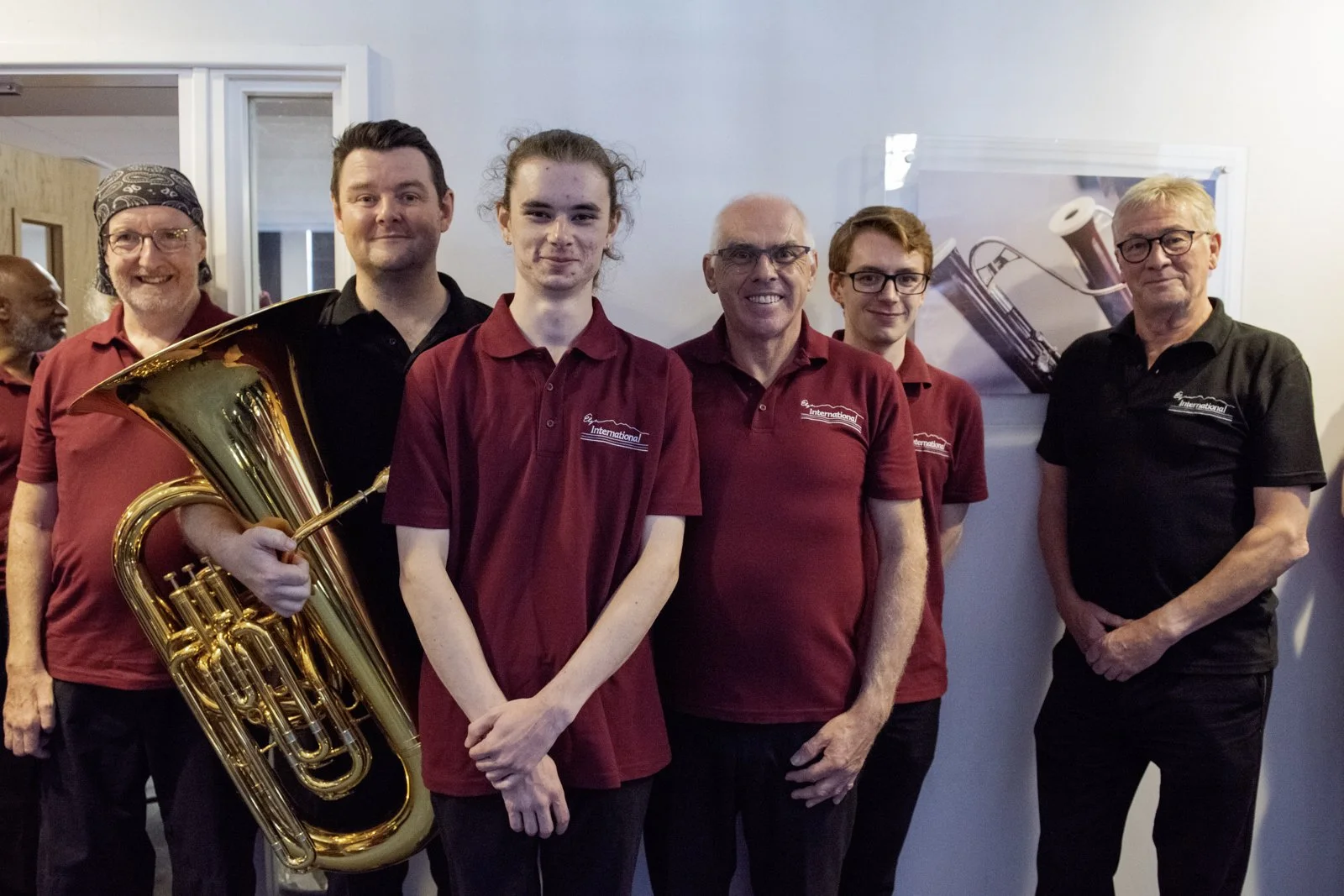 Group of six men standing indoors, dressed in red and black shirts, with one man holding a large brass musical instrument, possibly a euphonium, smiling and posing for the camera.