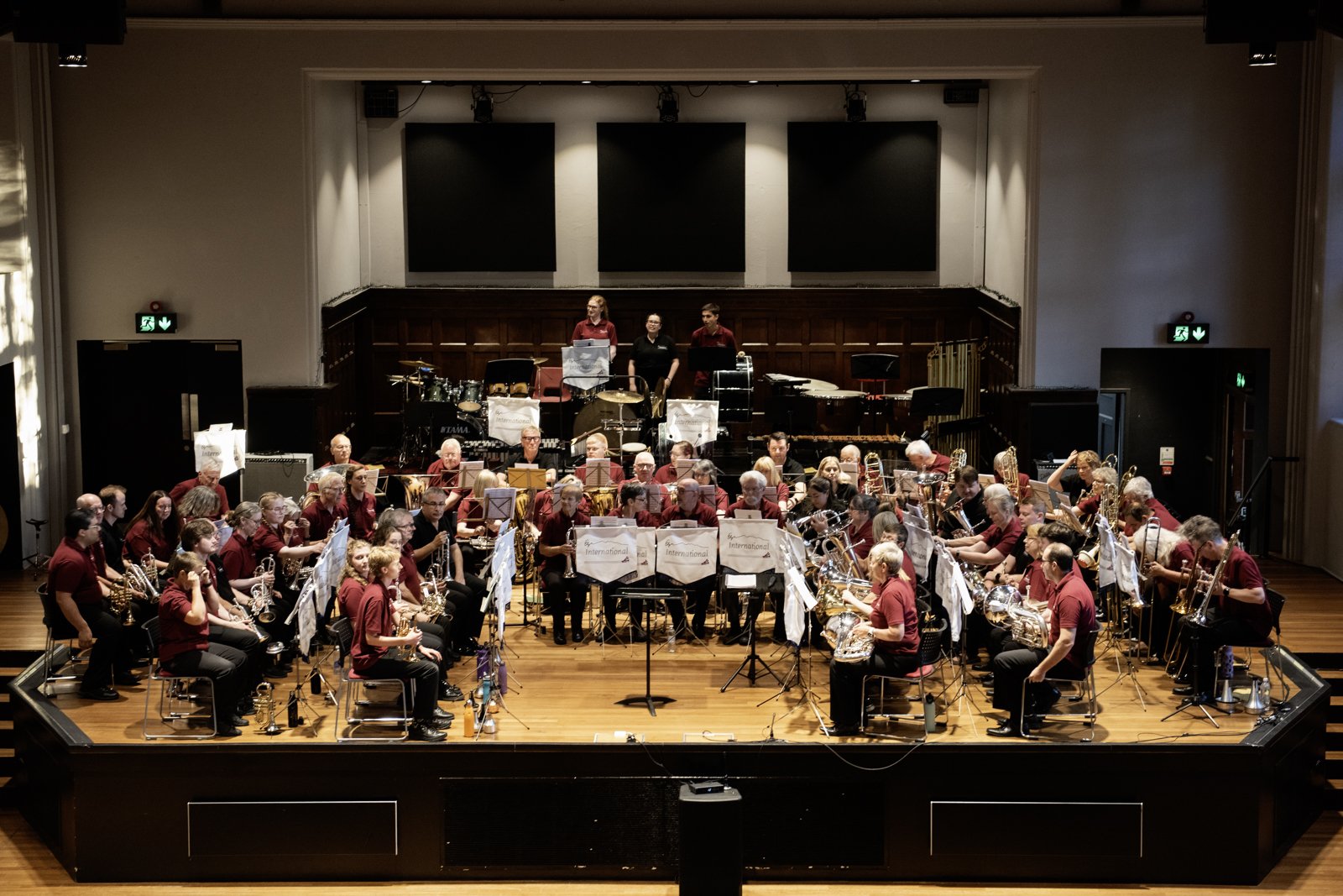 Symphony orchestra on stage in concert hall, with musicians in burgundy shirts, music stands, and various instruments, stage with wooden floor, large blank screens, and musicians standing behind, in front of an audience.