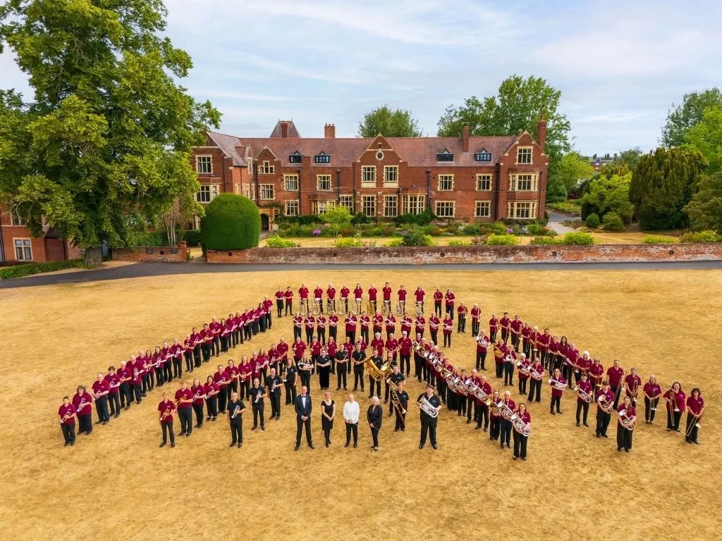A large group of musicians, part of a band, arranged in the shape of a heart on a dry, grassy field in front of a large red-brick building with a garden. The band members are wearing maroon shirts with black pants, and some hold their instruments.