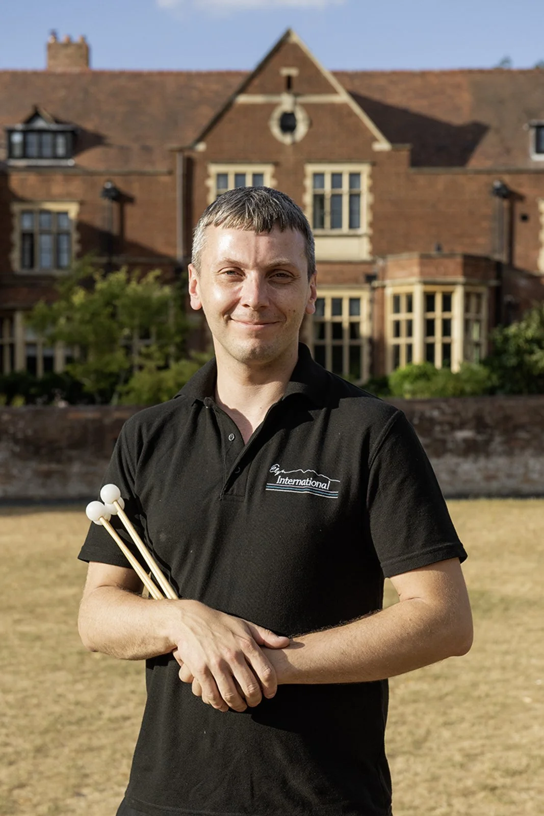 A man holding drumsticks standing outdoors in front of a large brick house with multiple windows and a well-maintained garden.
