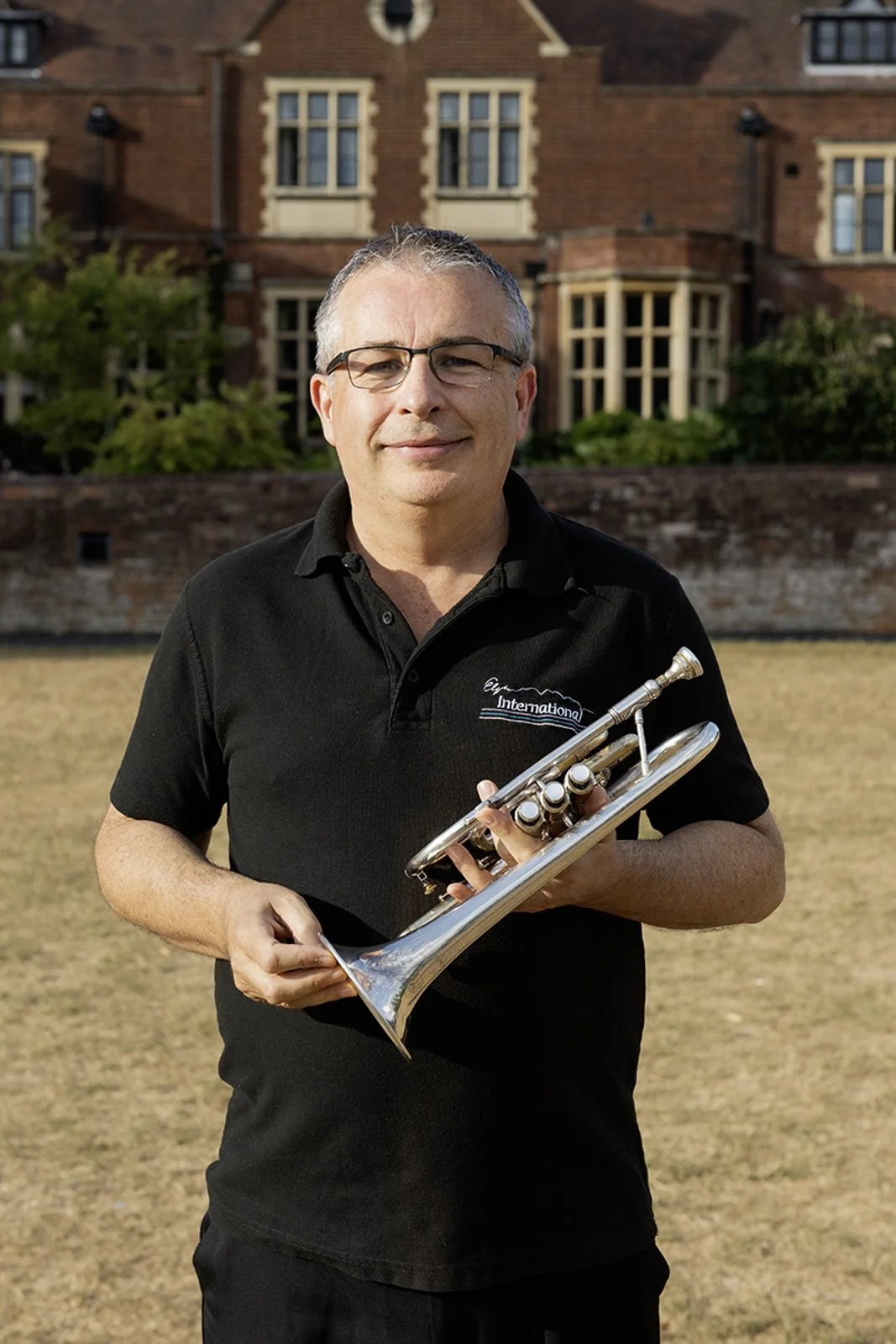 A man with glasses holding a trumpet in front of a brick house with multiple windows and green bushes.