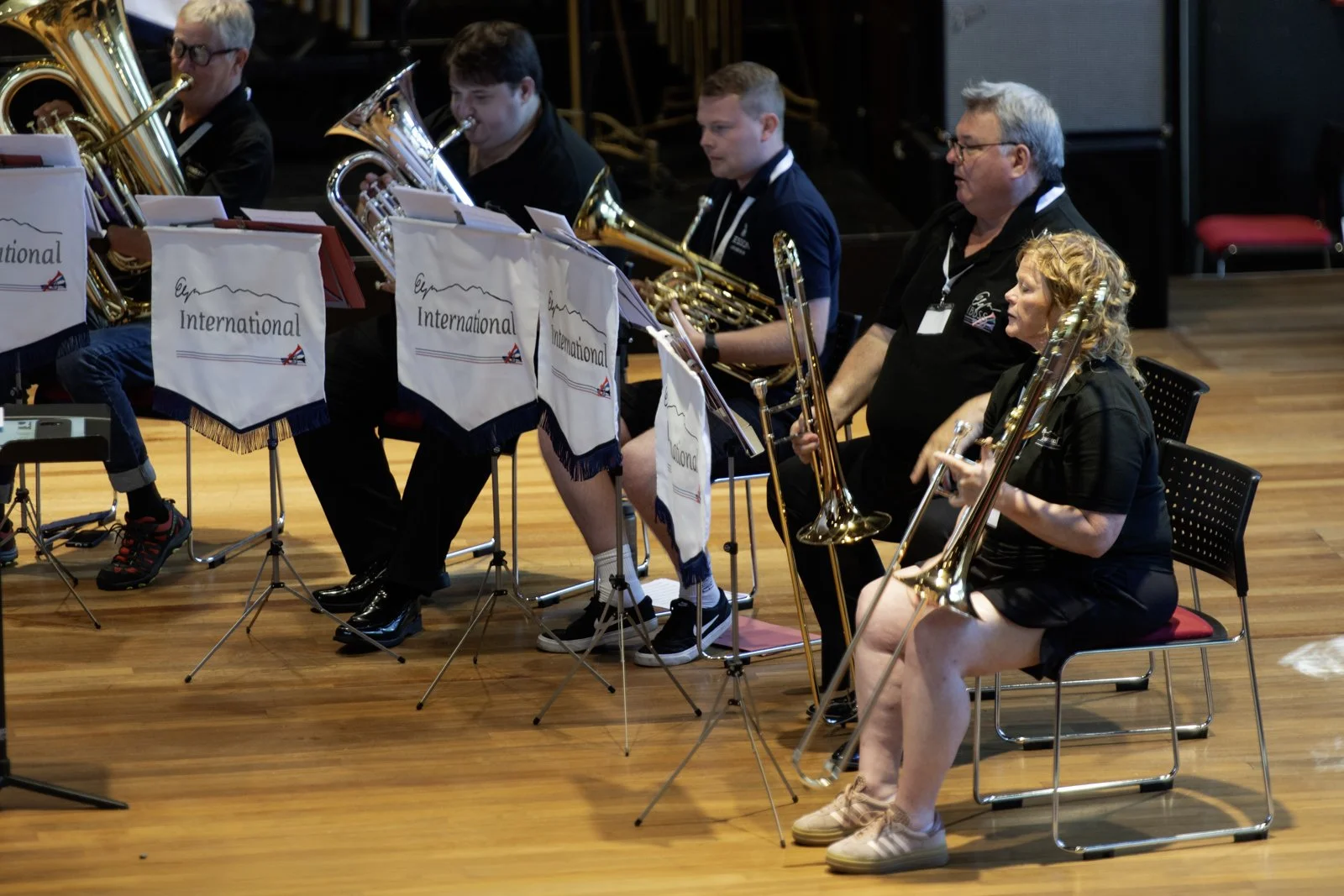 A group of musicians playing brass instruments during a performance. The musicians are seated on chairs on a wooden stage, with music stands in front of them that display banners reading 'International' with a mountain outline and a small British fla