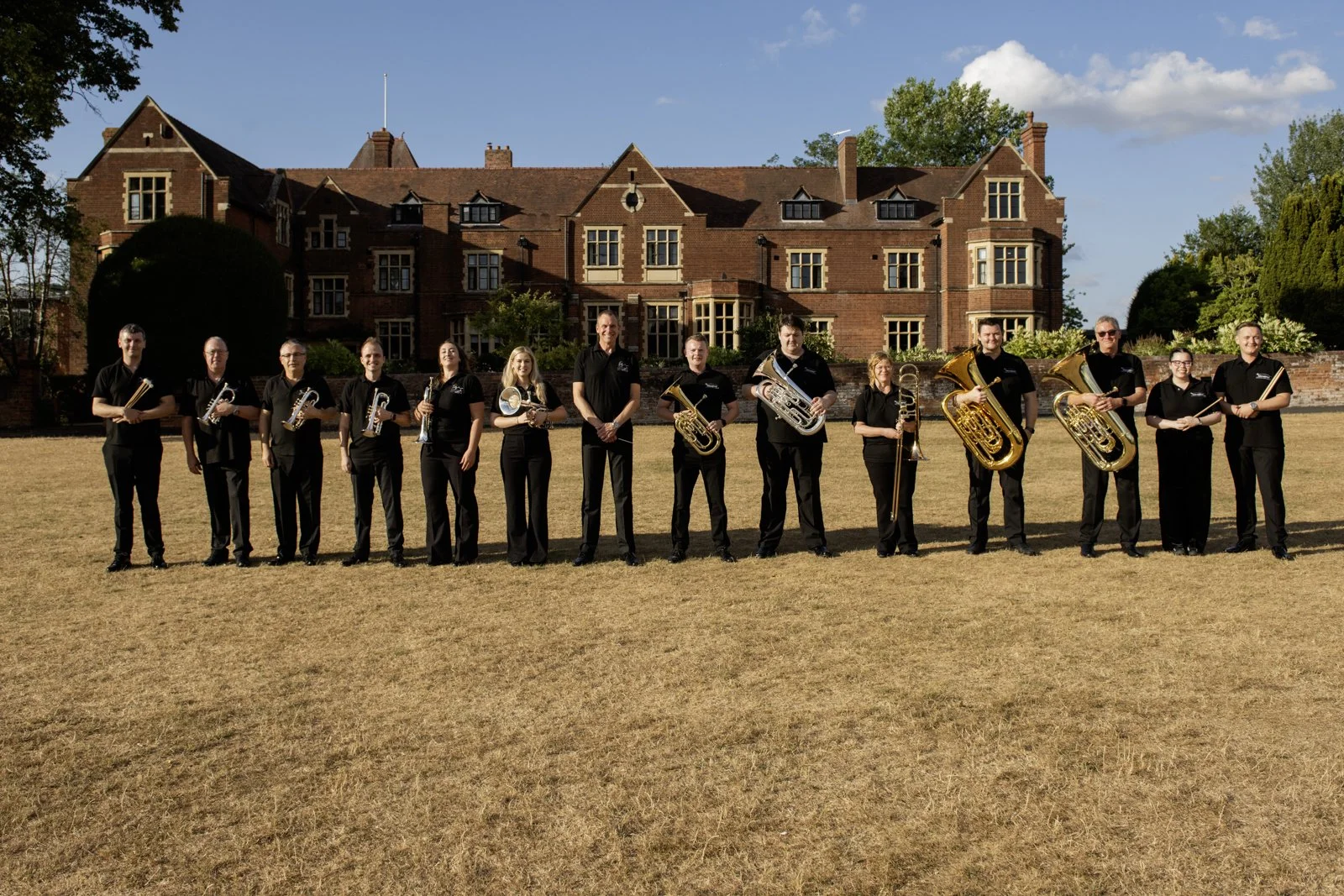 A group of 12 musicians standing outdoors on a field with a large brick building in the background. They are holding musical instruments including trumpets, a flute, a euphonium, and a tuba, and are dressed in black attire.