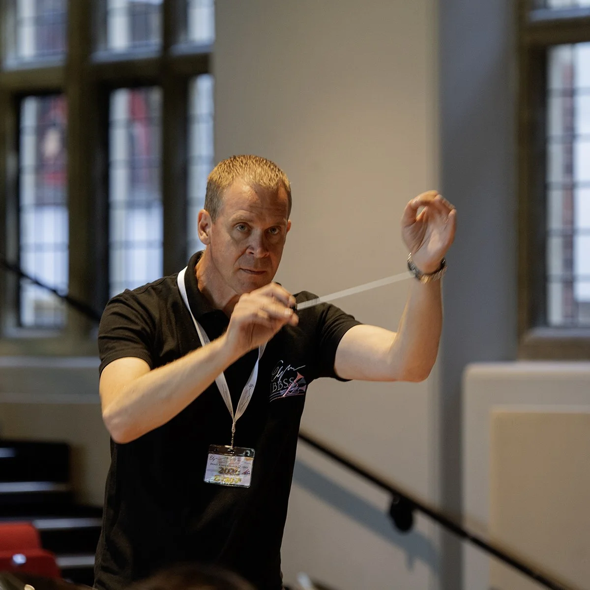 A man conducting an orchestra or choir, holding a baton in his right hand and guiding with his left, inside a room with large windows.