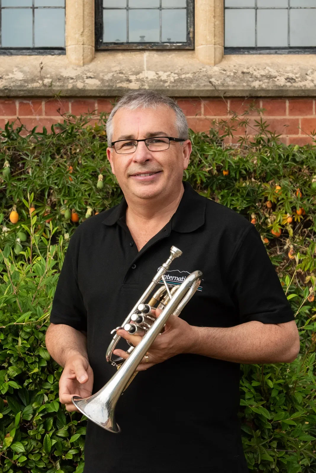 A man with gray hair, glasses, and a black collared shirt holding a silver trumpet, standing outdoors with green bushes and small orange fruits behind him.