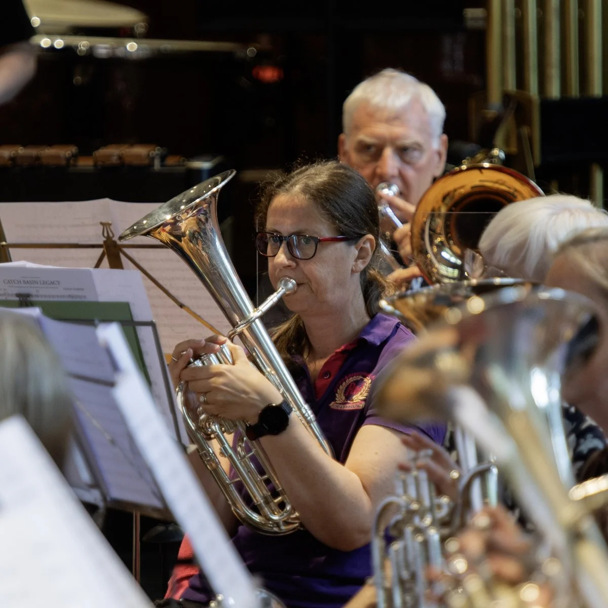 Woman with glasses playing a brass instrument in an orchestra, with other musicians and sheet music in the background.