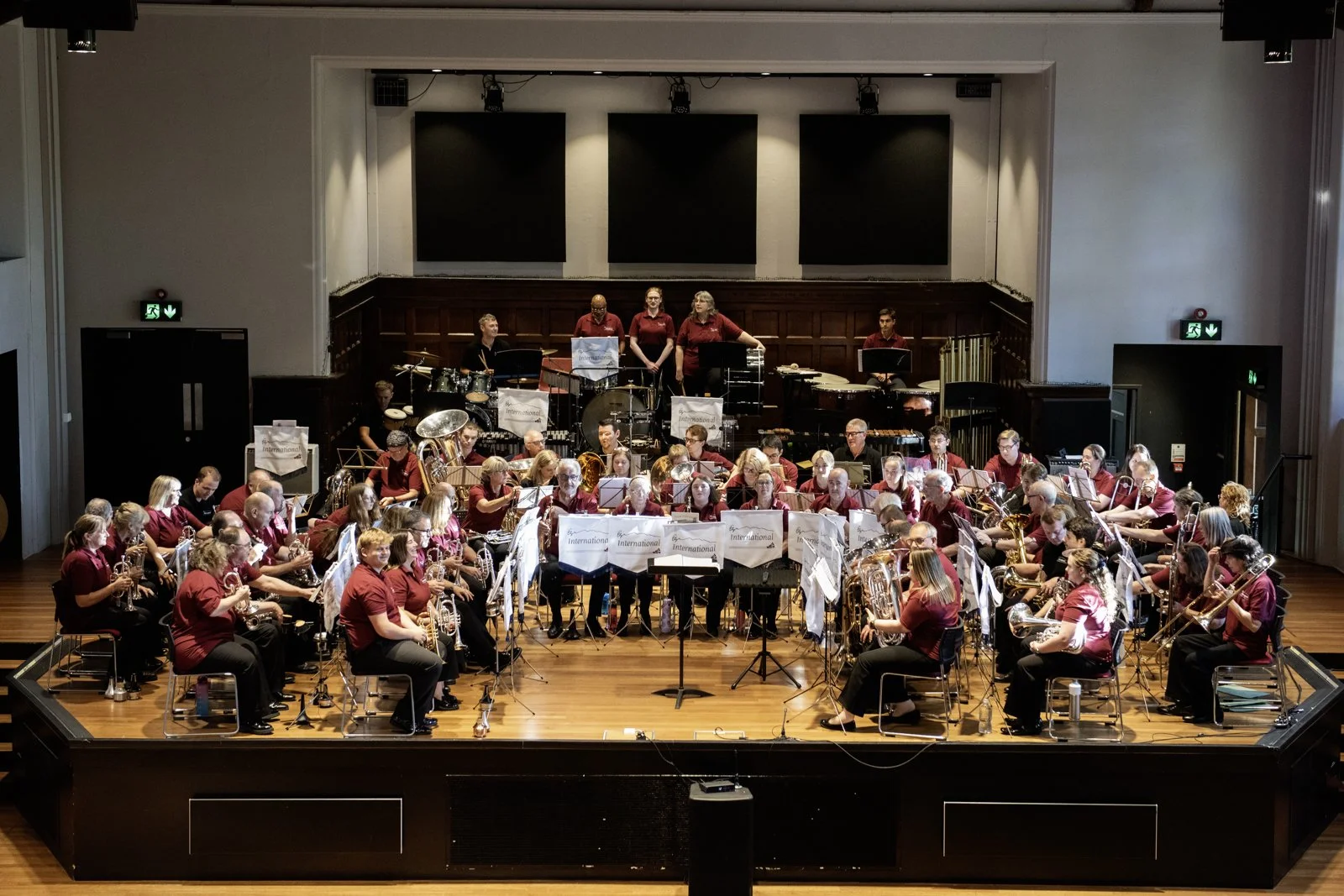 A large symphony orchestra performing on a wooden stage in a concert hall with music stands, various instruments, and musicians wearing matching maroon shirts.