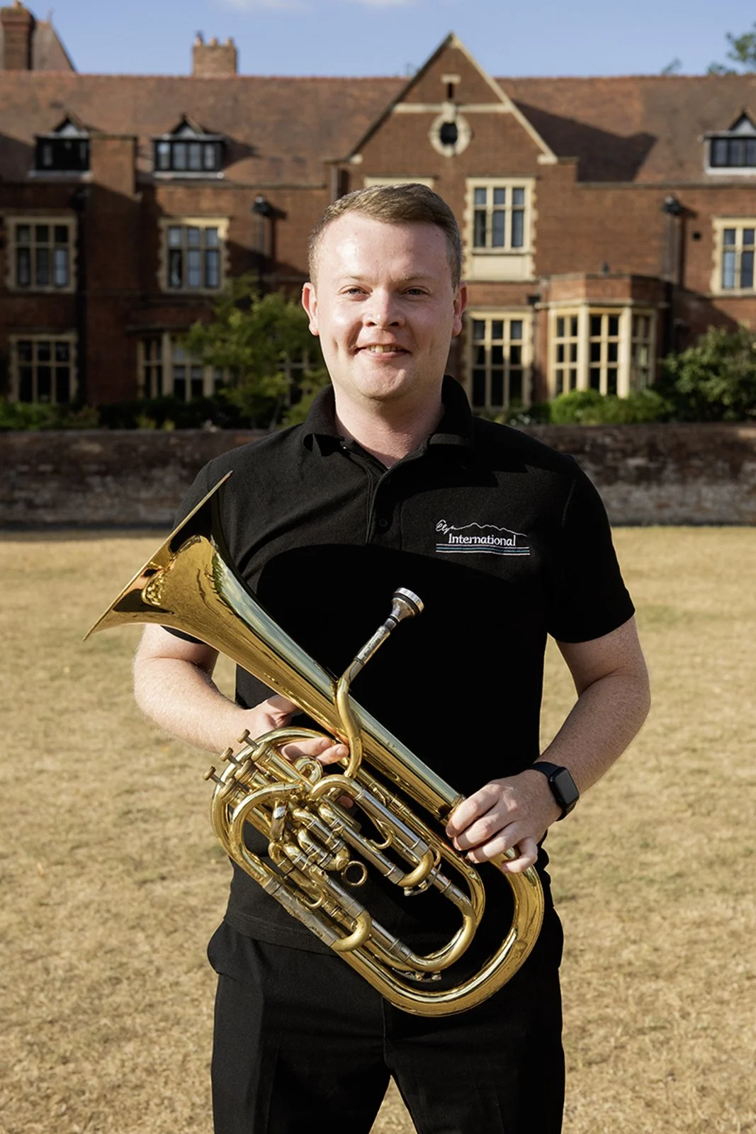 A young man standing outdoors on a sunny day holding a brass instrument, possibly a euphonium or baritone horn, in front of a large brick building with many windows and a well-kept garden.