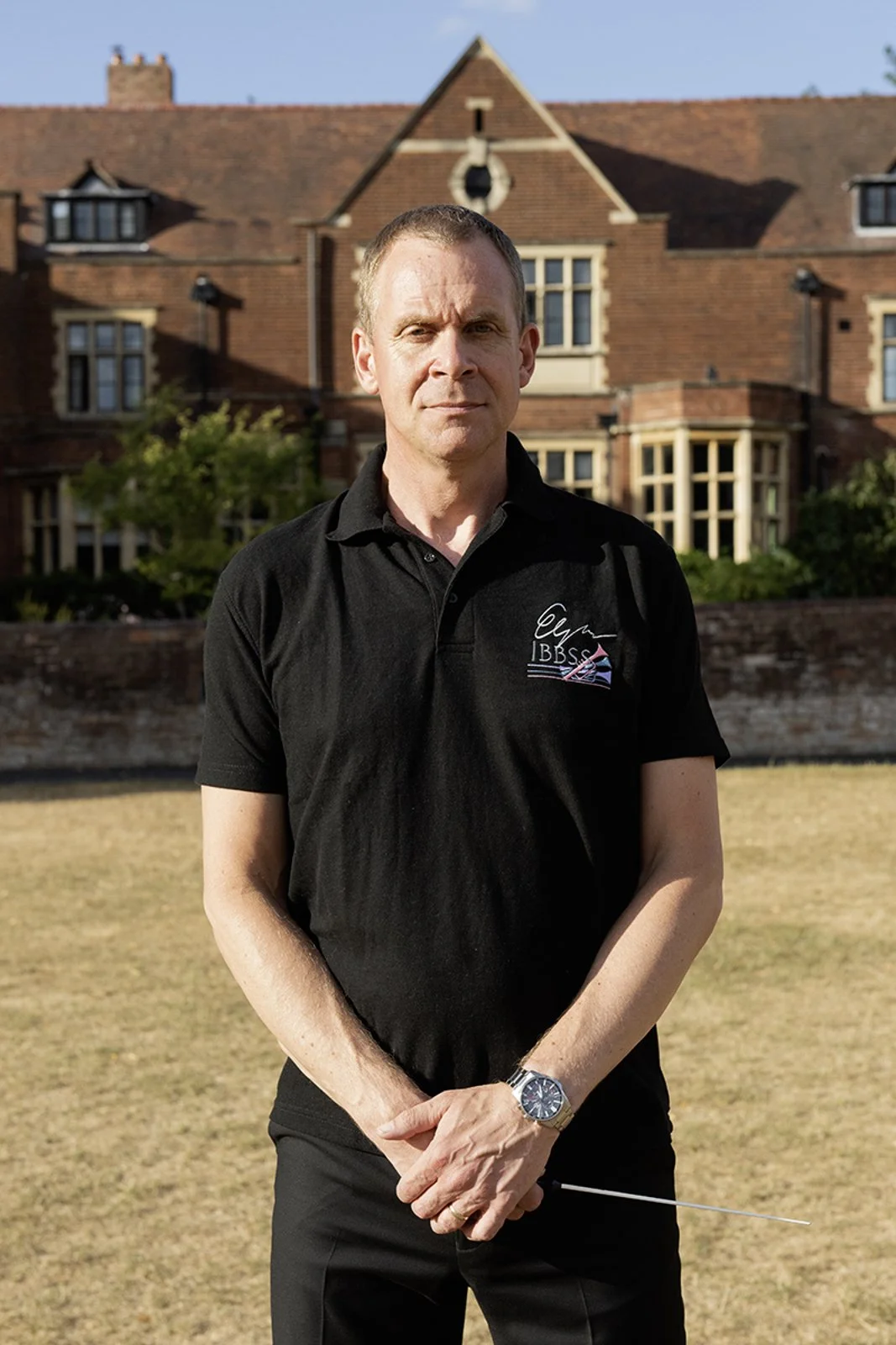 A man standing outdoors on a grassy area in front of a brick house with a pointed roof and multiple windows, wearing a black polo shirt and a silver wristwatch.