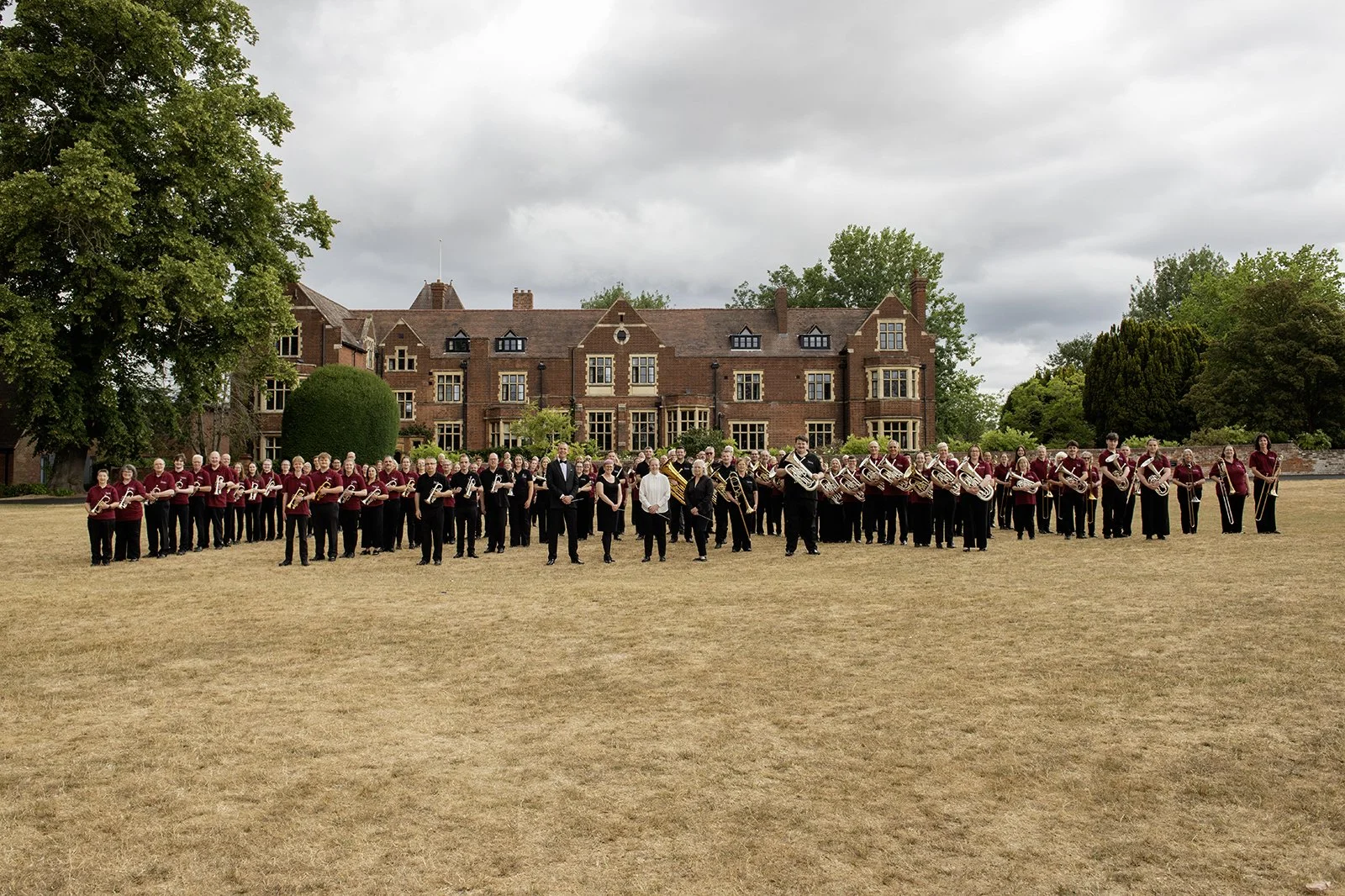 A large group of people in formal and coordinated outfits, holding musical instruments, standing on a dry grass field in front of a large brick building with trees around. The sky is cloudy.