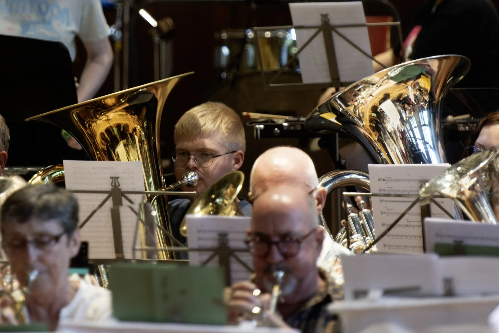Members of an orchestra playing instruments, including tubas and clarinets, with sheet music on stands in a venue with warm lighting.