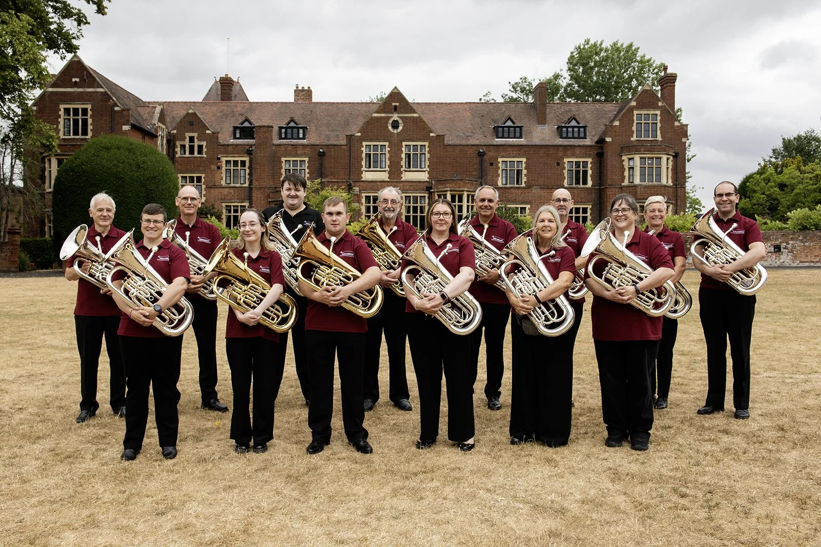 A group of 13 people, all holding French horns, standing outdoors on a grassy area in front of a large brick mansion with trees in the background.