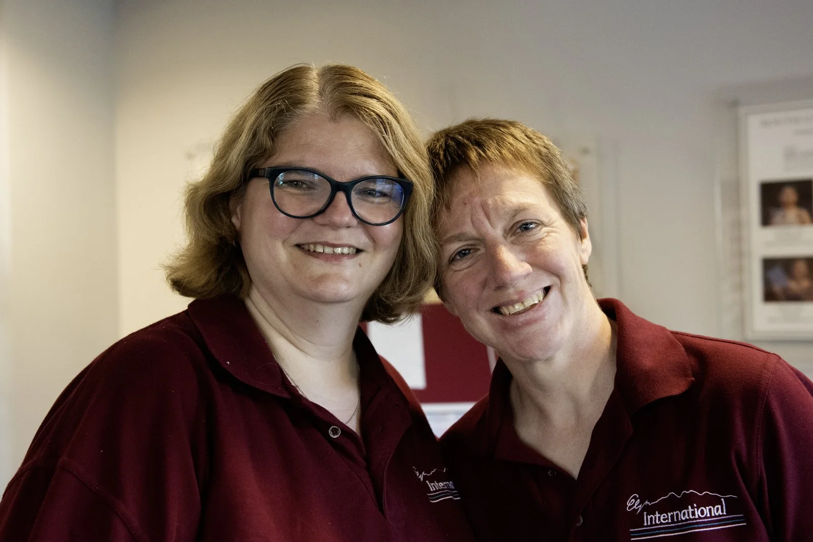 Two women smiling, standing close together indoors, both wearing maroon shirts with white text and logo, with photos on the wall in the background.