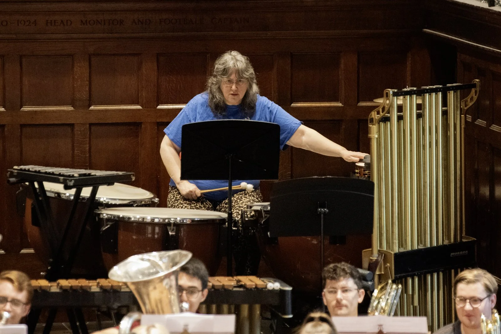 A woman with gray hair, wearing a blue shirt and leopard print pants, conducting an orchestra with percussion instruments in front of a wooden panel background.