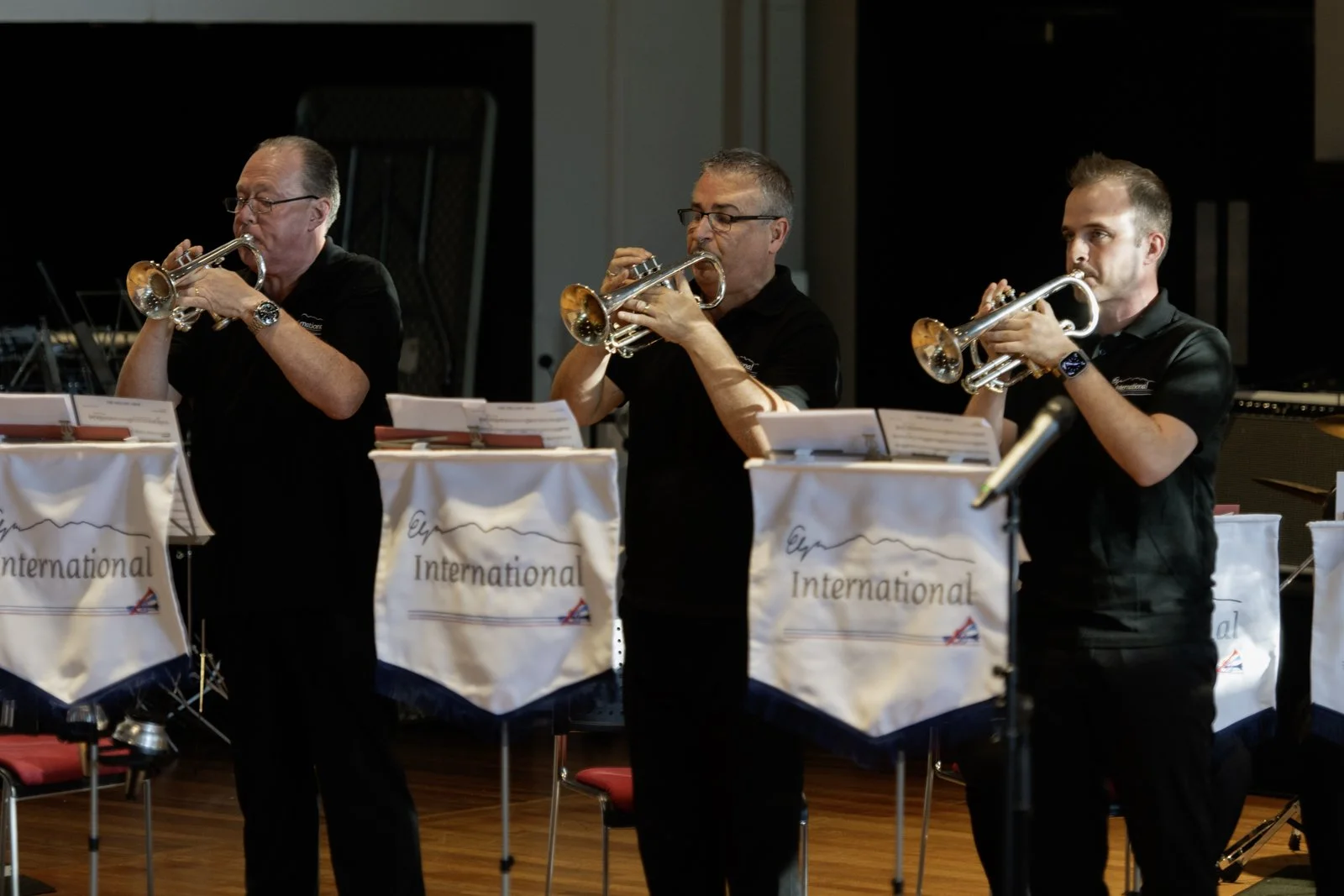 Three musicians playing trumpets on stage, wearing black shirts, in front of music stands with banners that read "International."