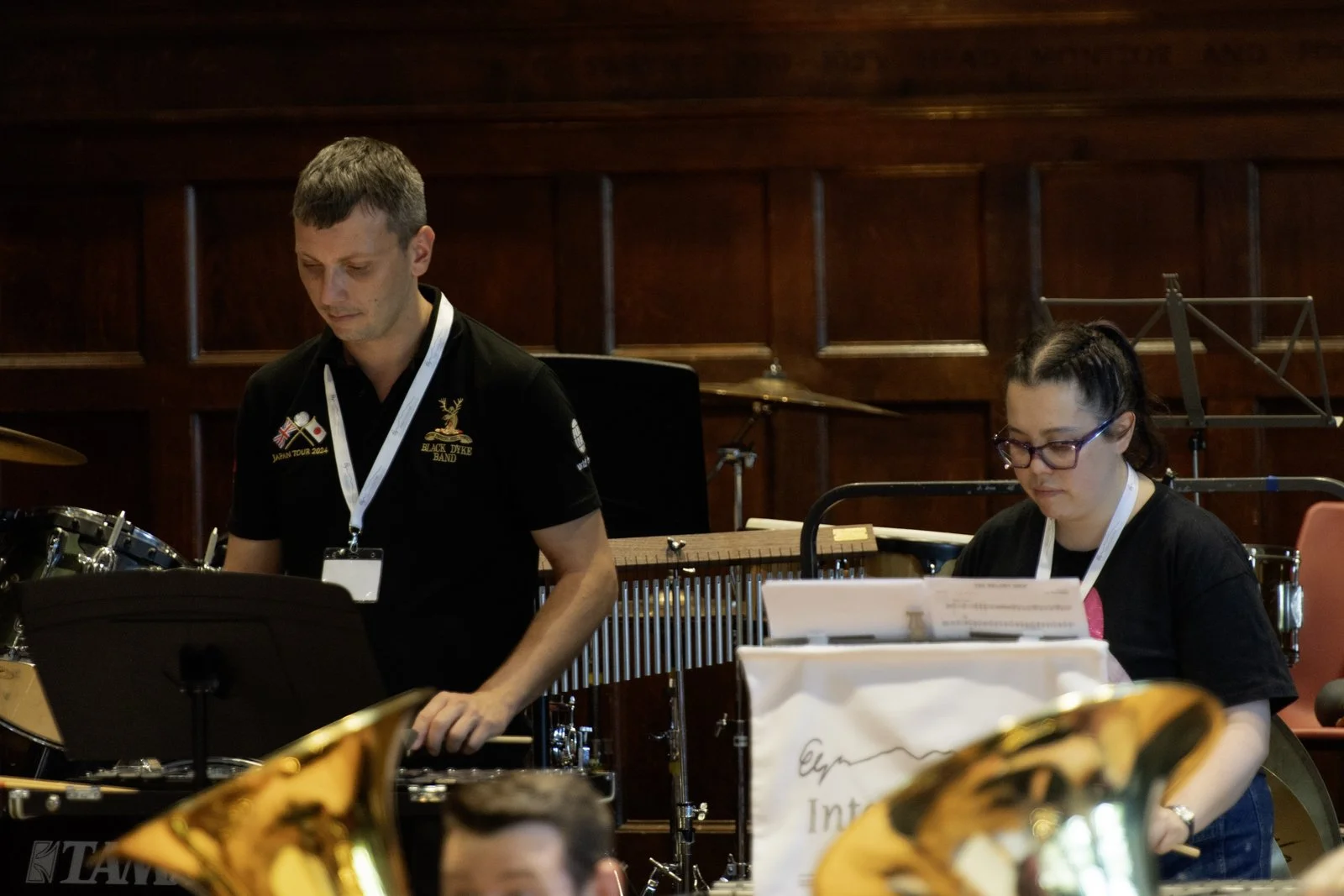 Two musicians, a man and a woman, are preparing musical instruments in a wooden-paneled room. The man on the left is looking down at a music stand, and the woman on the right is adjusting her music sheets, both are wearing black shirts with music eve