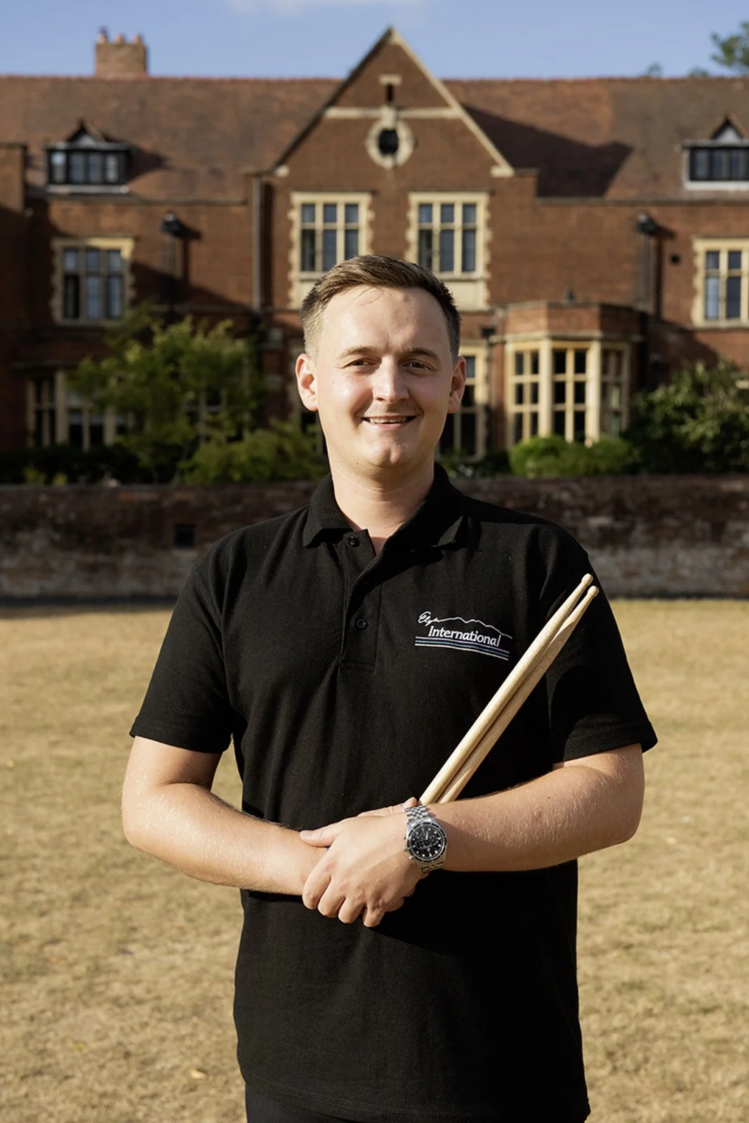 A young man holding drumsticks outdoors in front of a large brick house with multiple windows and a gabled roof, smiling at the camera.