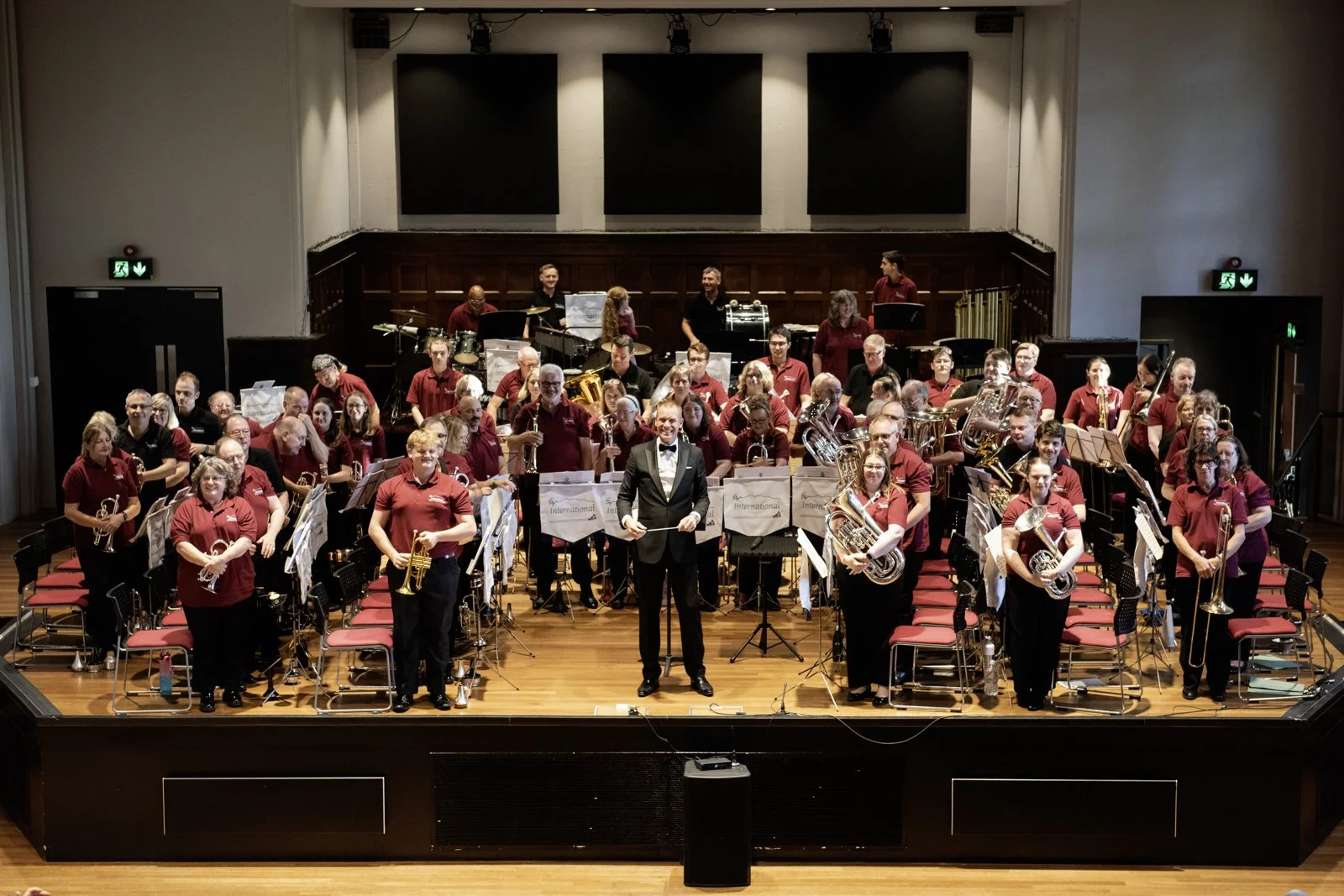 A large orchestra composed of musicians and a conductor on stage in a concert hall, with some members holding brass, woodwind and percussion instruments, and a person in a tuxedo standing at the center.