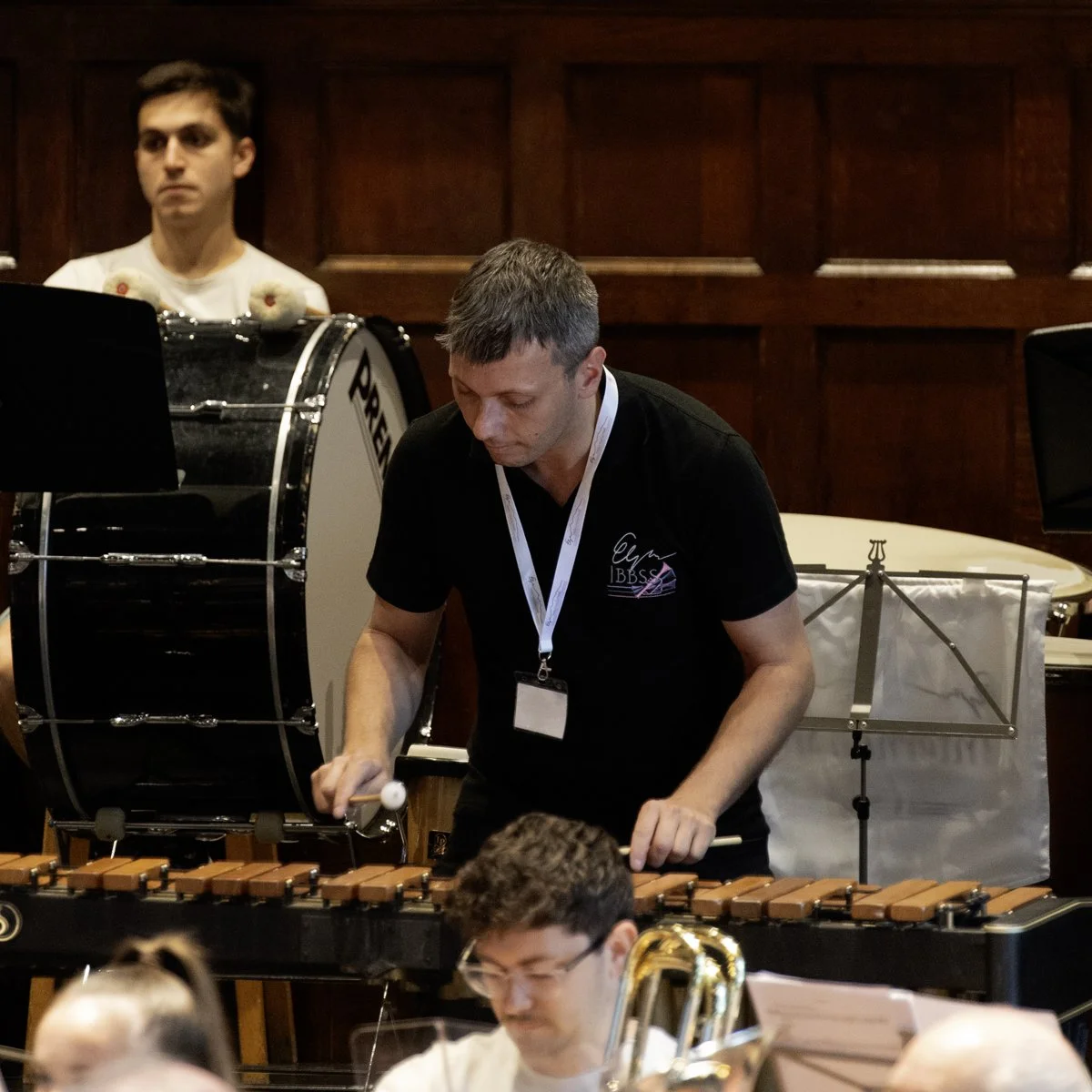 Two male musicians performing in an indoor setting with wooden paneling. One is playing a marimba, and the other is standing behind a bass drum, with several other musicians and instruments partially visible in the foreground.