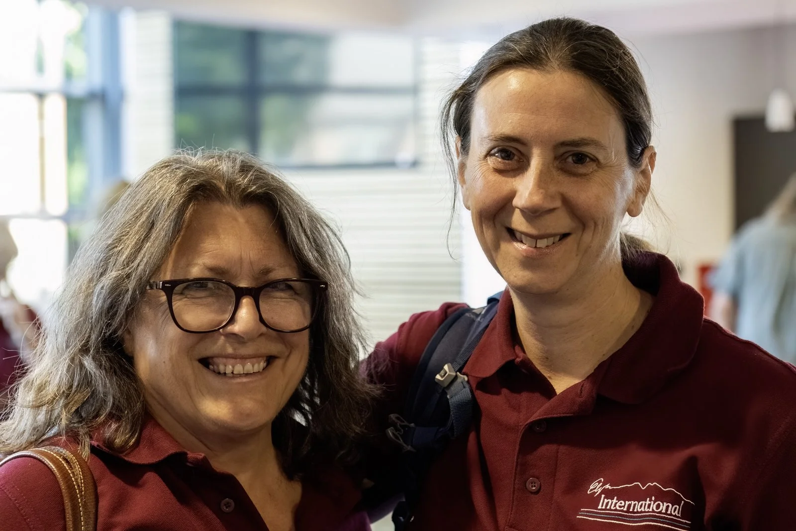 A smiling woman wearing glasses and a maroon shirt standing next to a smiling woman in a maroon polo shirt with a backpack, inside a well-lit building.