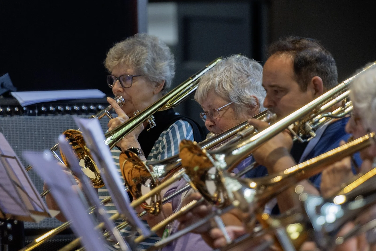 A group of people playing brass instruments, likely trombones, in an indoor setting, with music stands and sheet music visible.