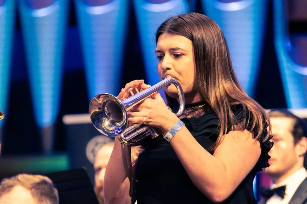Woman playing a silver trumpet at a formal event, wearing a black dress and a wristband, with other audience members in the background.