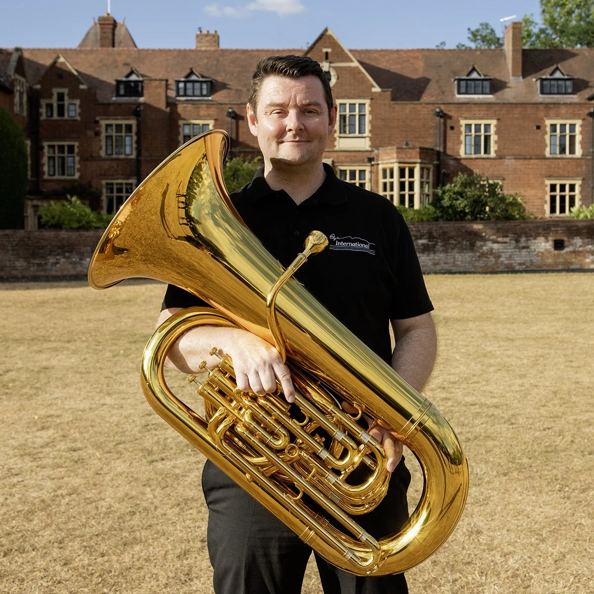 A man standing outdoors on a grassy area, holding a large brass tuba. He is wearing a black polo shirt with a logo, and there are residential brick buildings in the background. Sunny weather with clear skies.