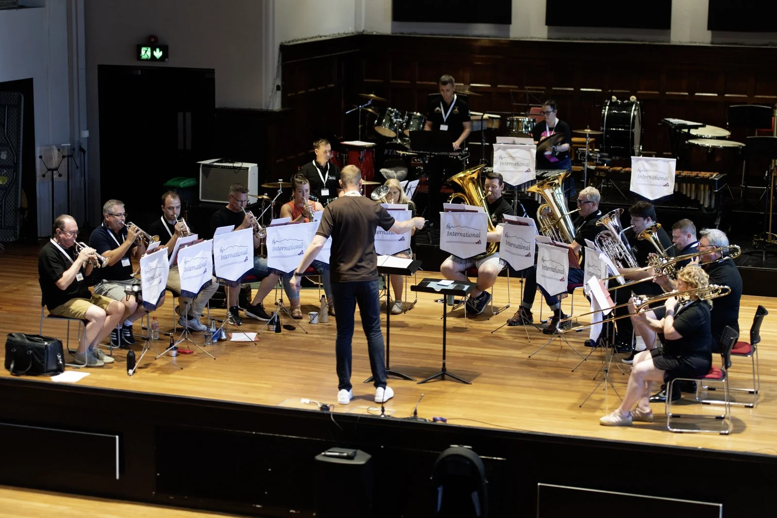 A large group of musicians playing brass and woodwind instruments during a rehearsal or performance, led by a conductor standing in front of them on a wooden stage.