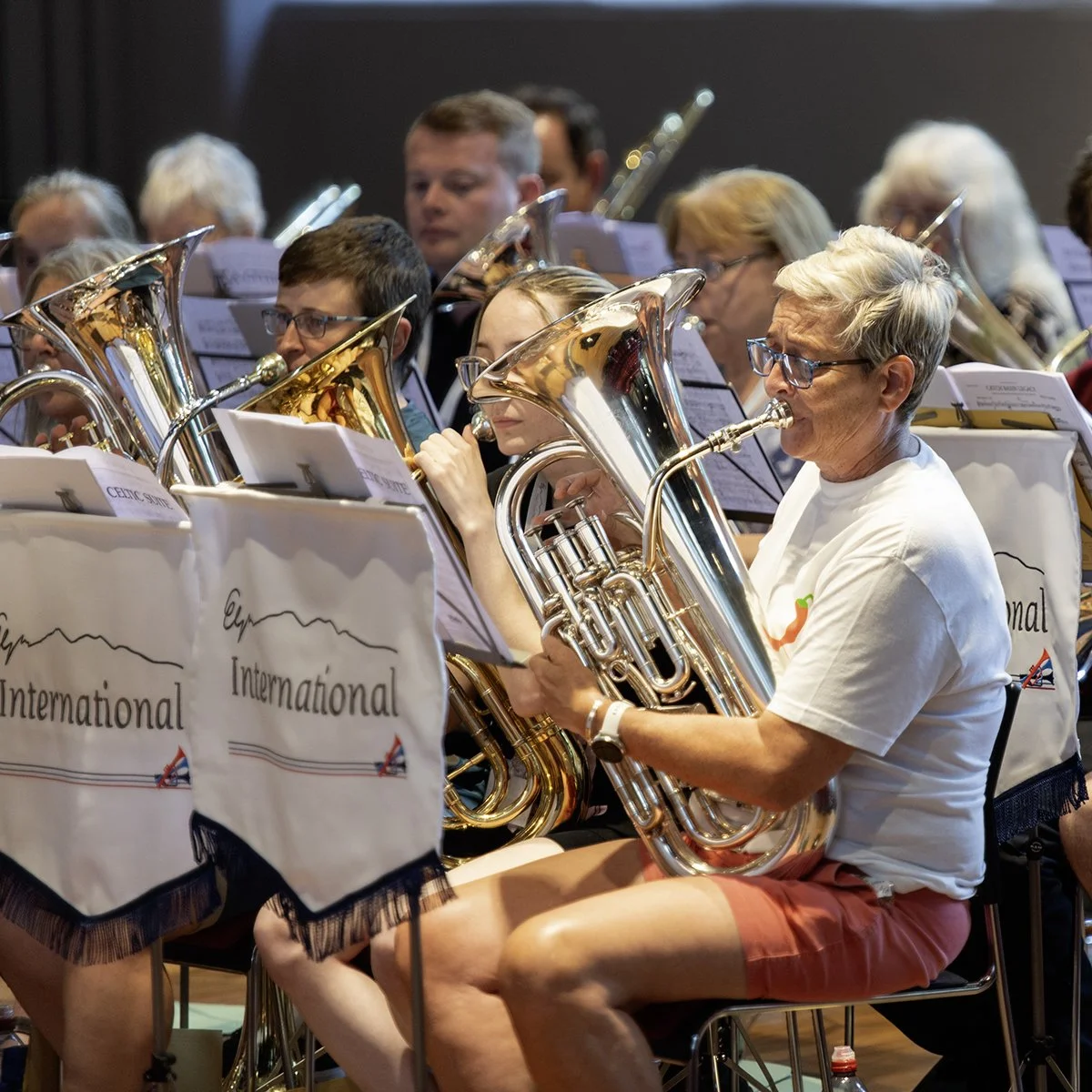 An orchestra performing, with members playing brass instruments like tubas, under sheet music stands labeled 'International'.
