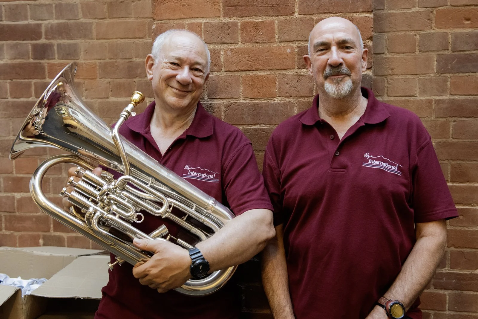 Two men standing in front of a brick wall, both wearing maroon polo shirts with a logo that says 'Egypt International.' The man on the left is holding a shiny silver tuba, smiling, and has white hair. The man on the right is bald with a gray beard, s