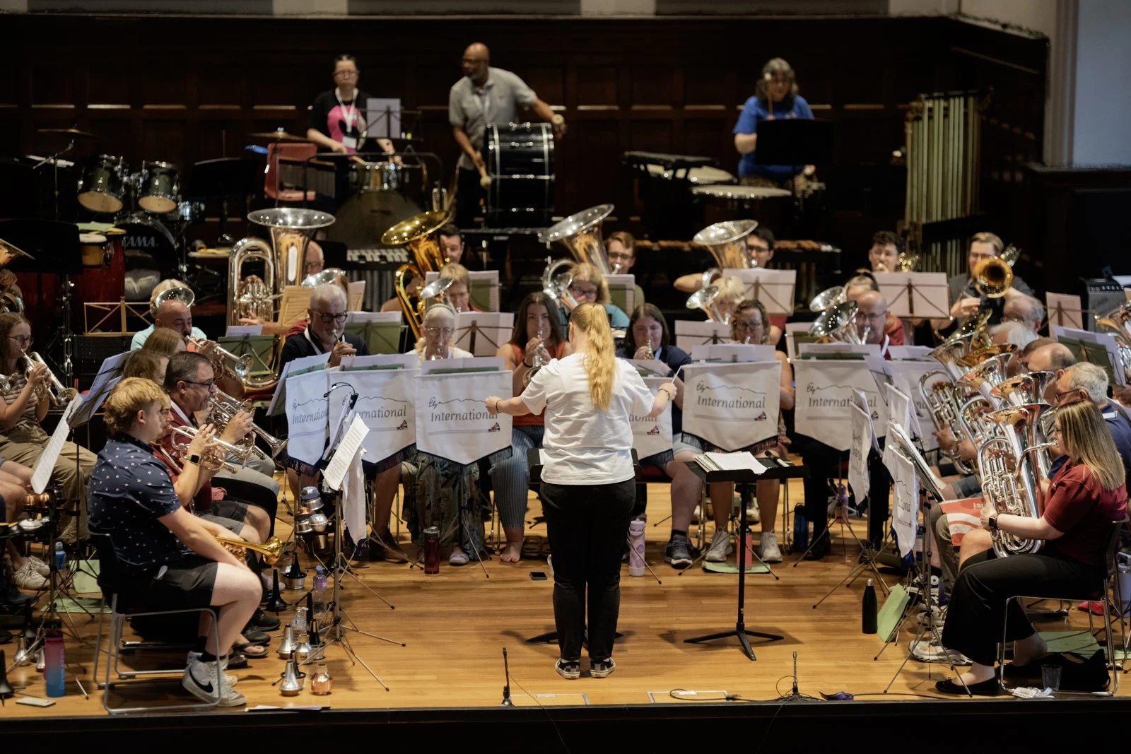 Orchestra rehearsal with musicians playing various instruments on a stage, conducted by a woman with long red hair. The backdrop includes drums and other percussion instruments.